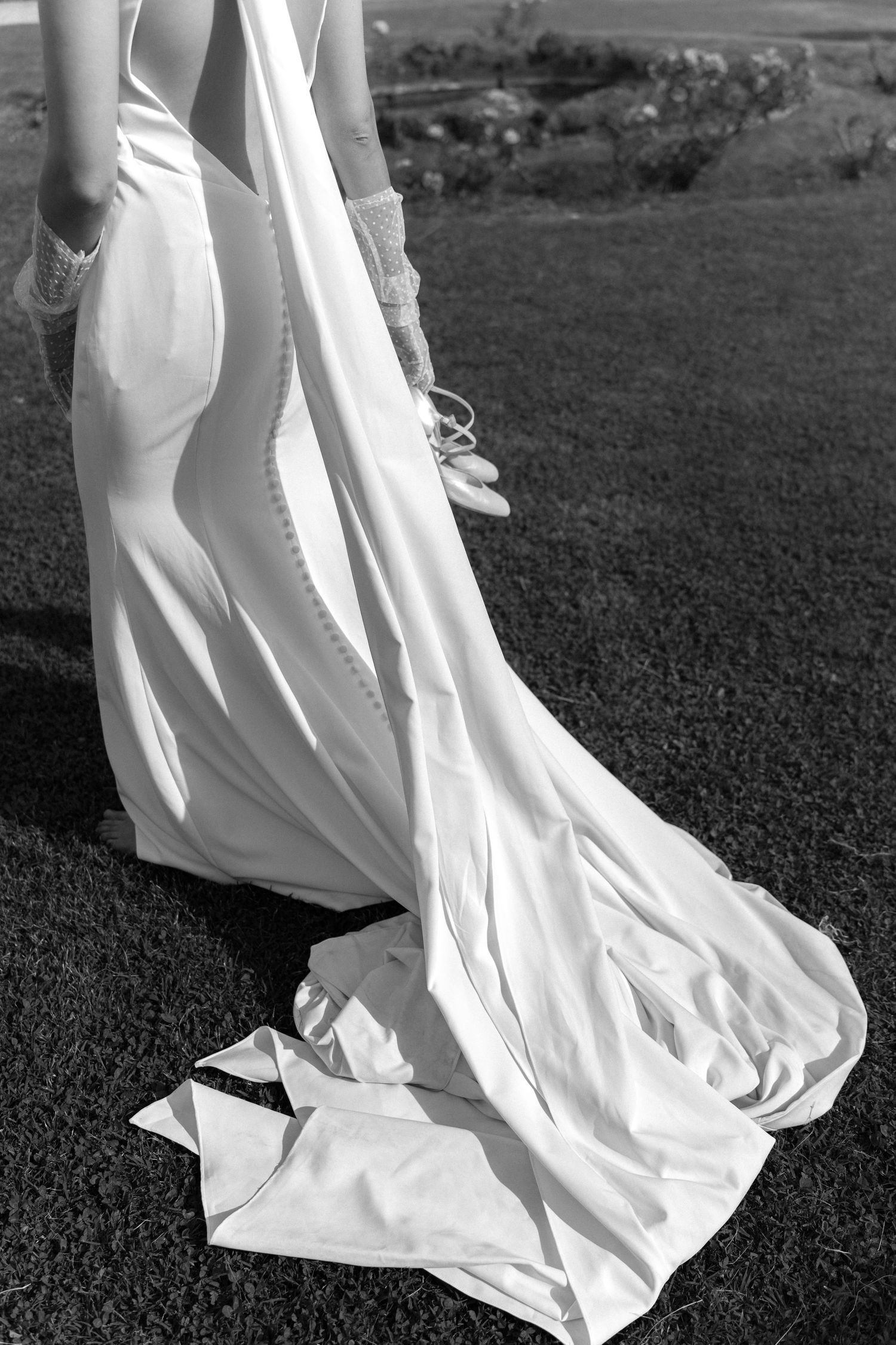 A flowing white wedding dress with a long train trails across green grass in a black and white photograph. Villa Mosconi Bertani