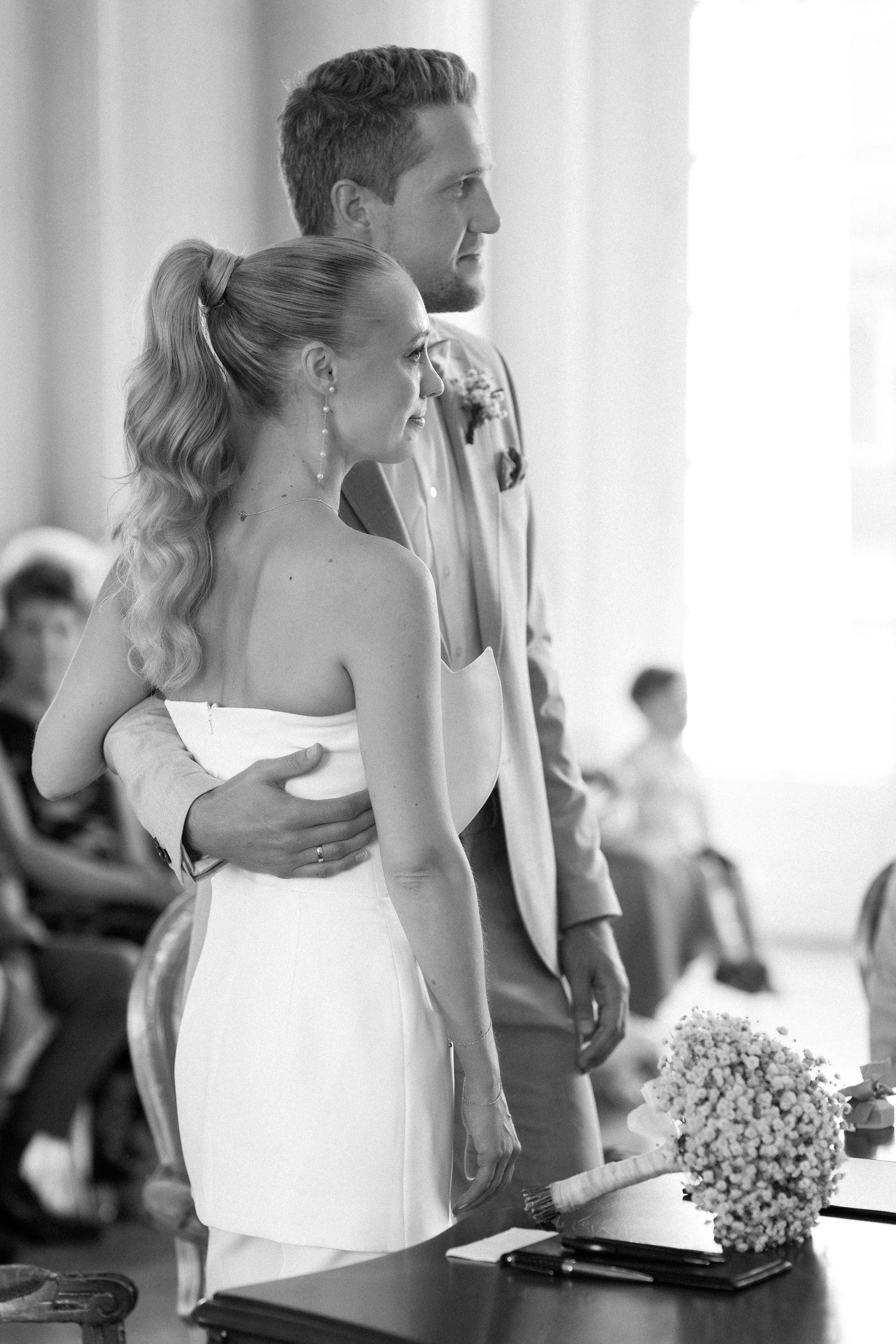 A couple stands together at a wedding ceremony in a white room with a small floral bouquet. Schloss Solitude