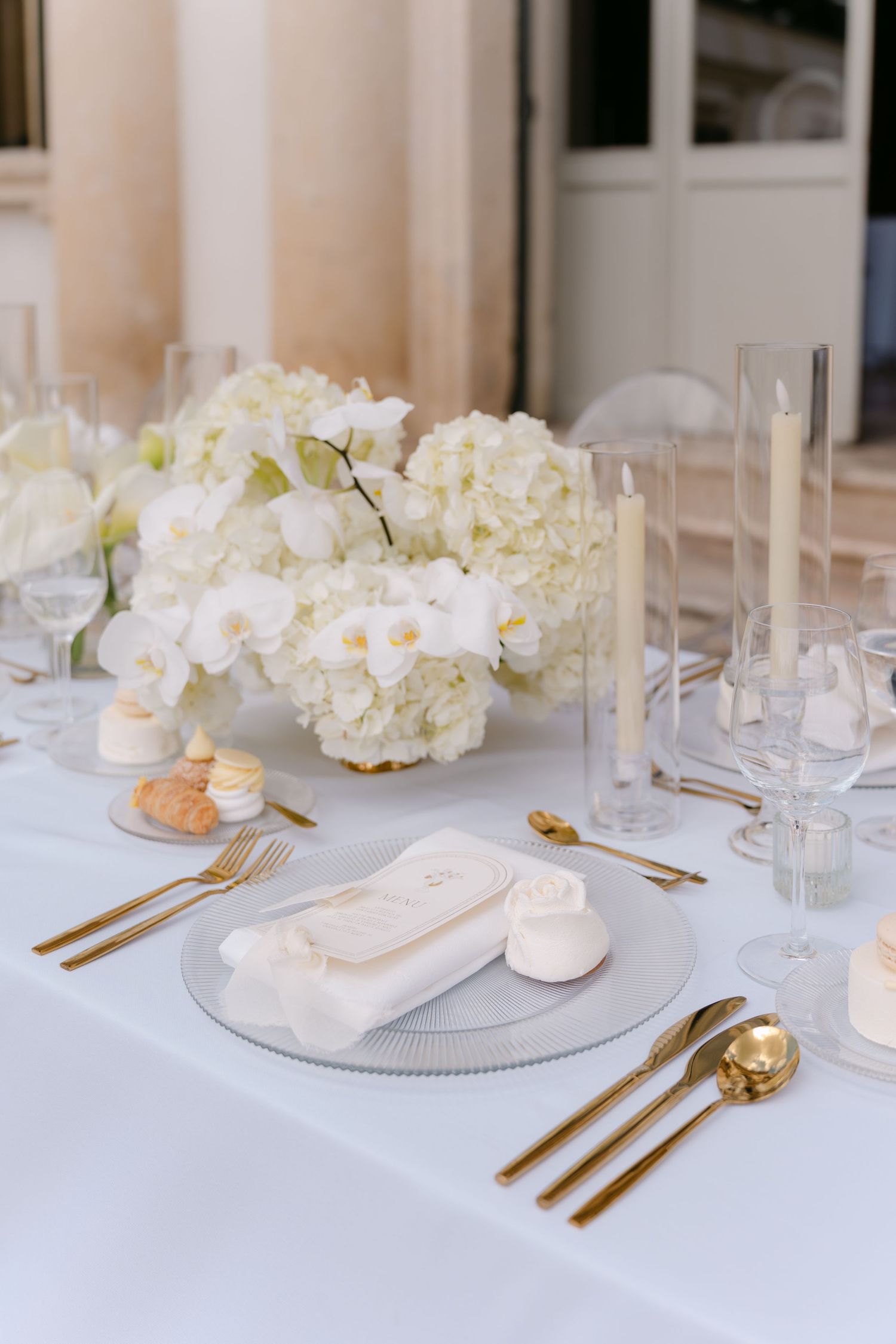 Elegant white and gold place settings with hydrangea centerpieces on a crisp white tablecloth at a formal event. Villa Mosconi Bertani
