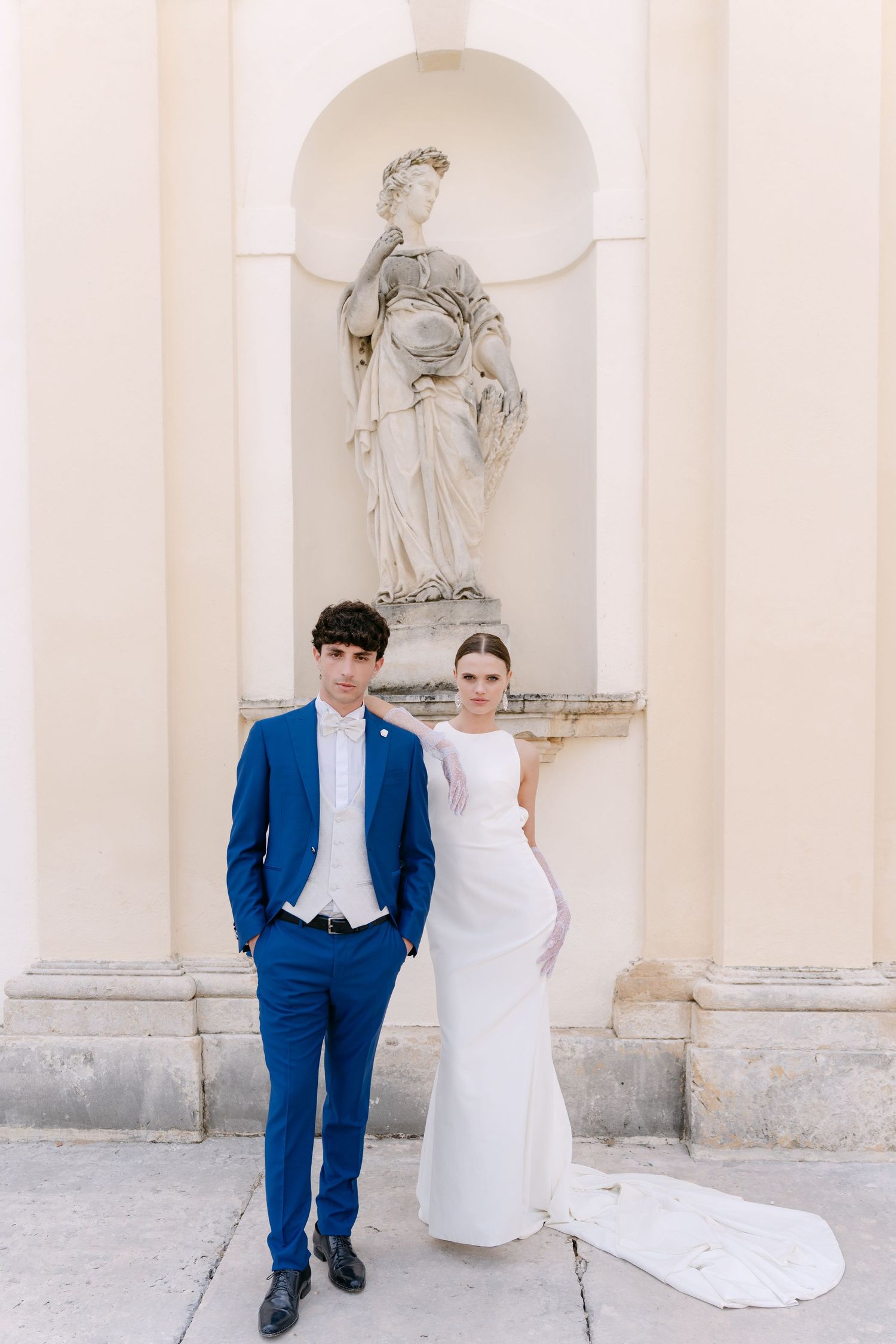Elegant wedding couple poses in front of white classical columns with decorative statues. Villa Mosconi Bertani
