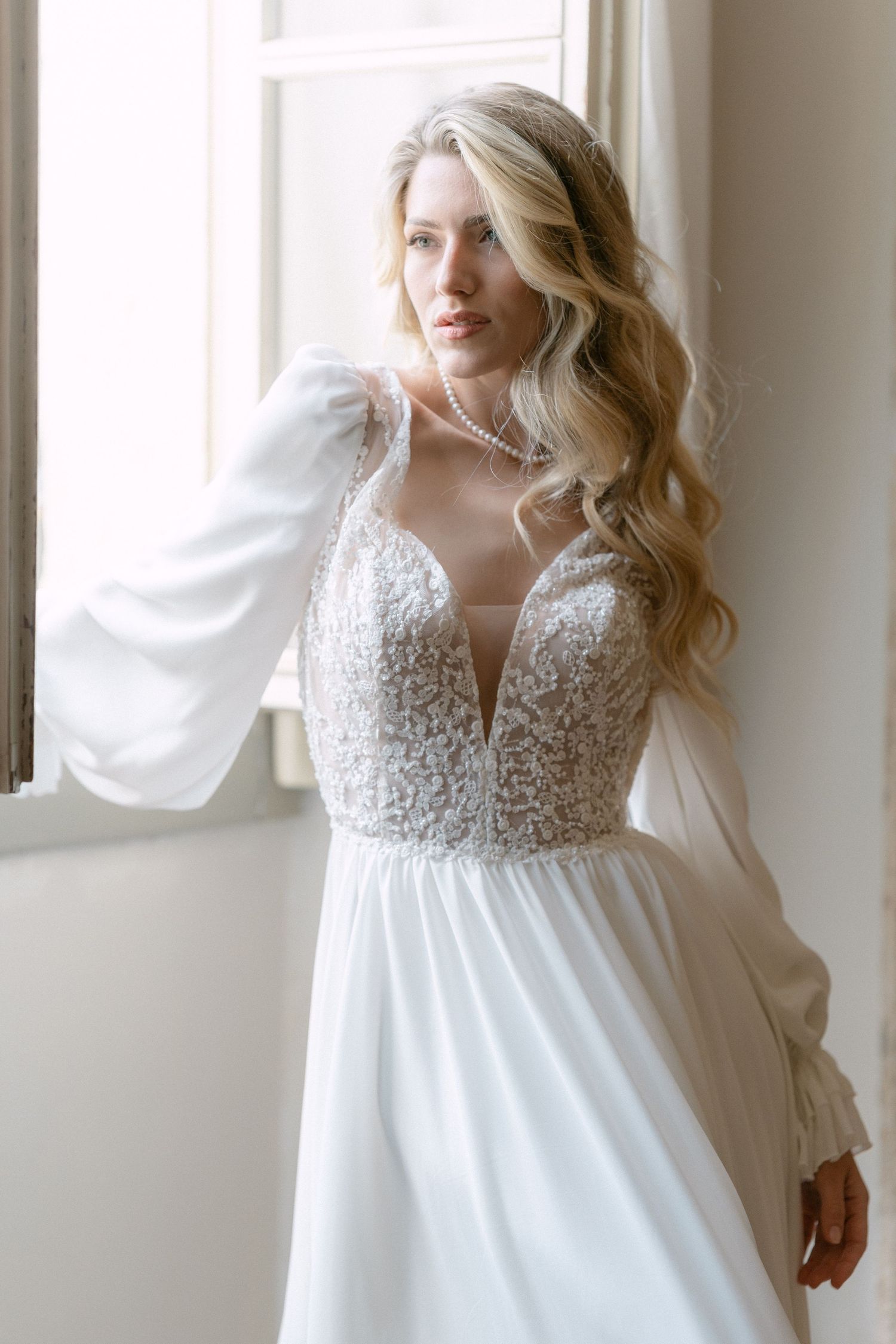 Elegant wedding dress with lace bodice, plunging neckline, sheer bishop sleeves and flowing white skirt by window. Wedding Verona