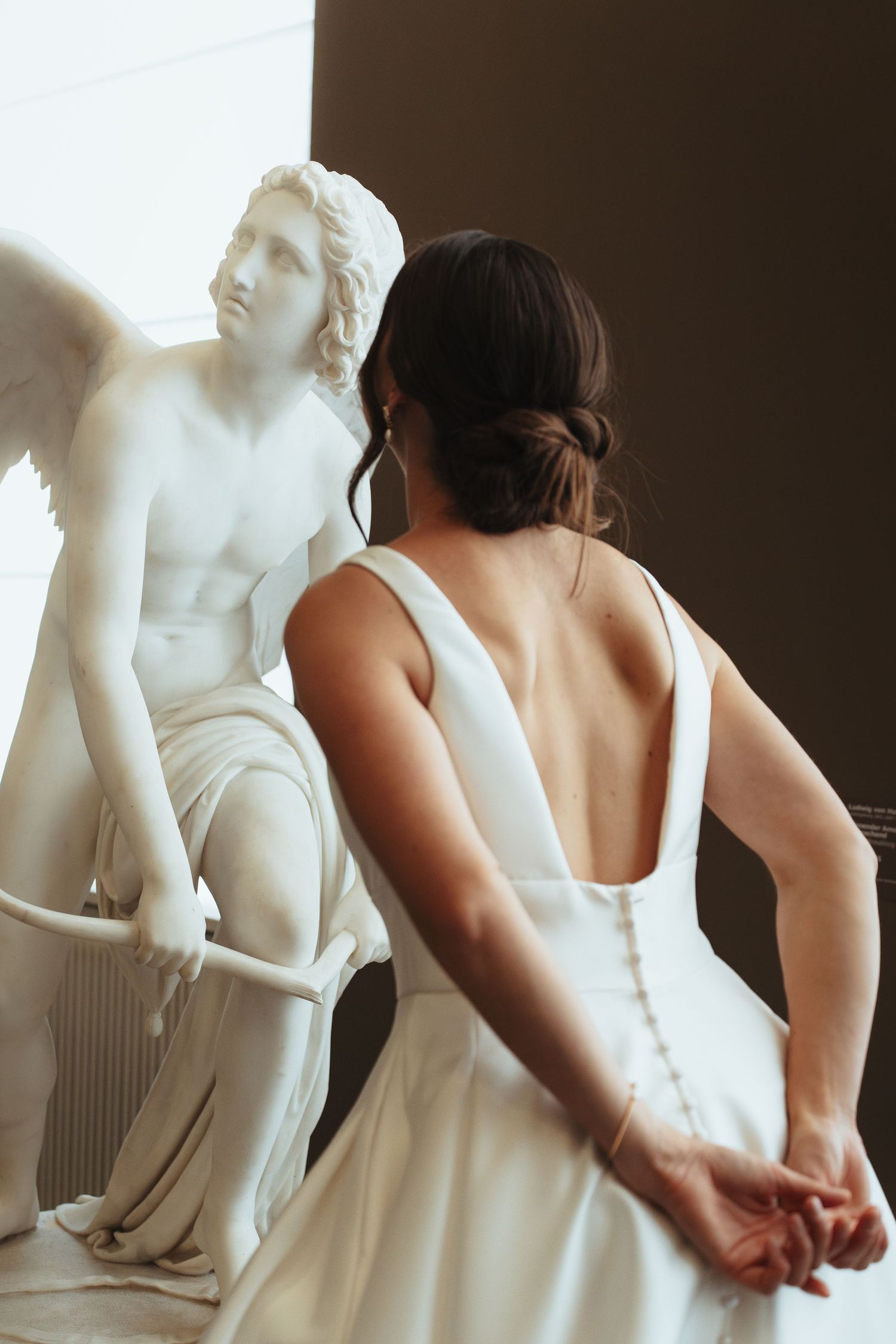 Woman in white wedding dress stands next to classical marble statue in an artistic bridal portrait.