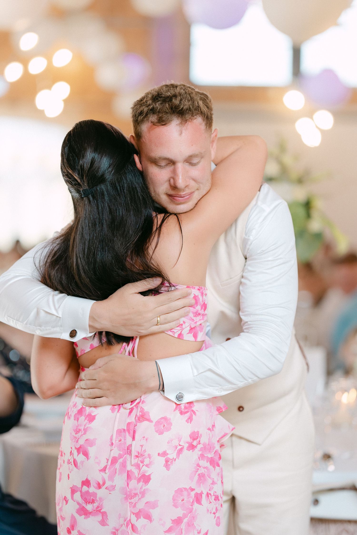 A couple shares an intimate dance moment at an event, with the woman wearing a pink floral dress and man in light suit.