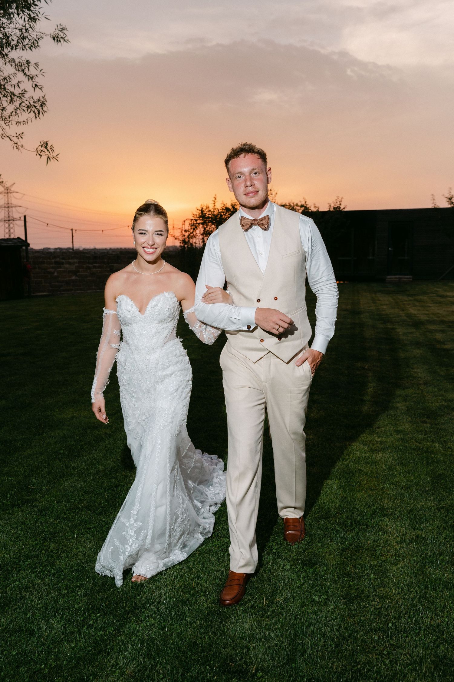A couple in wedding attire walks together on a grassy field at sunset during their special day.