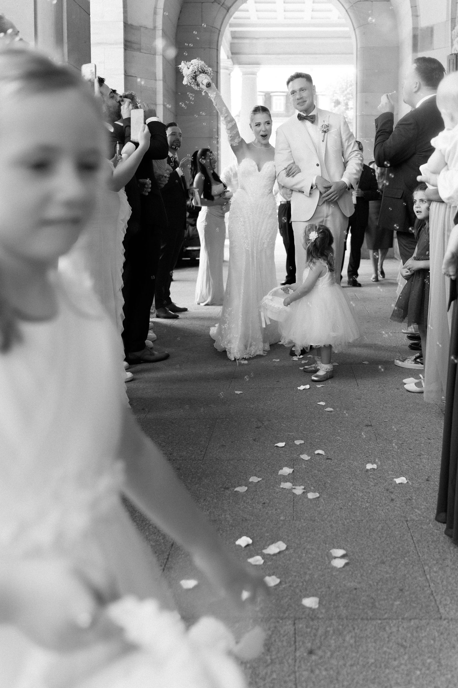 A couple walks on a petal-strewn aisle while wedding guests look on in a church hallway.