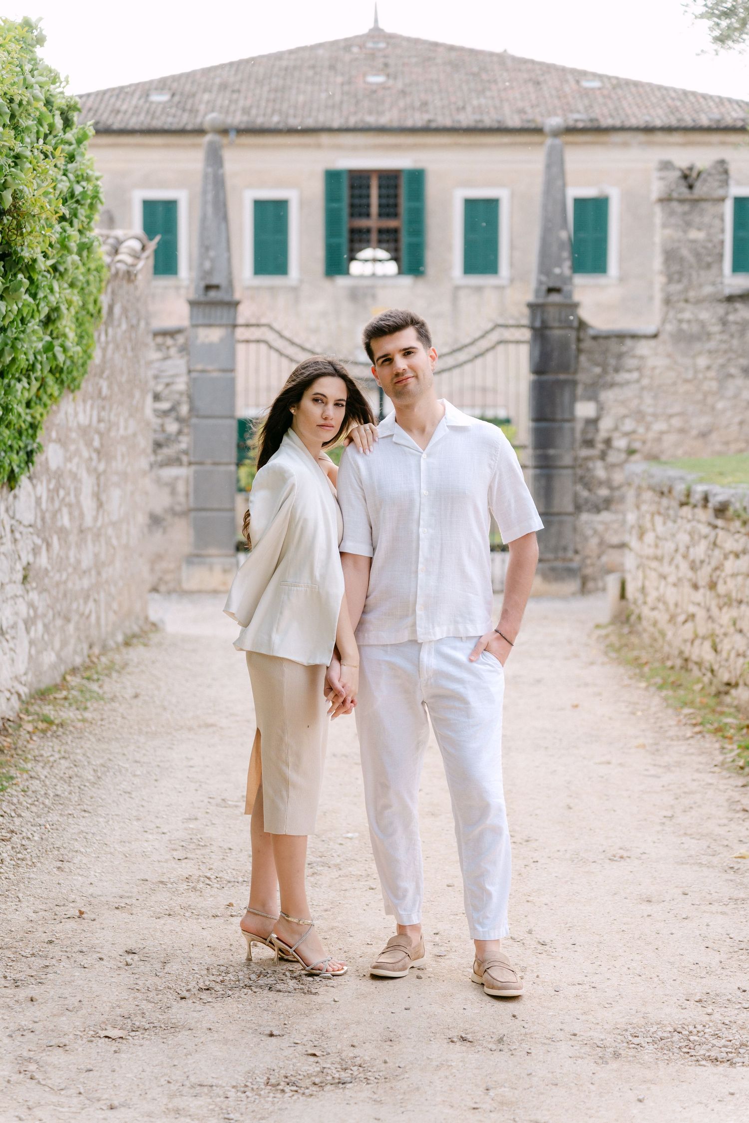 Couple in coordinated white outfits walking along a rustic stone path near historic buildings.