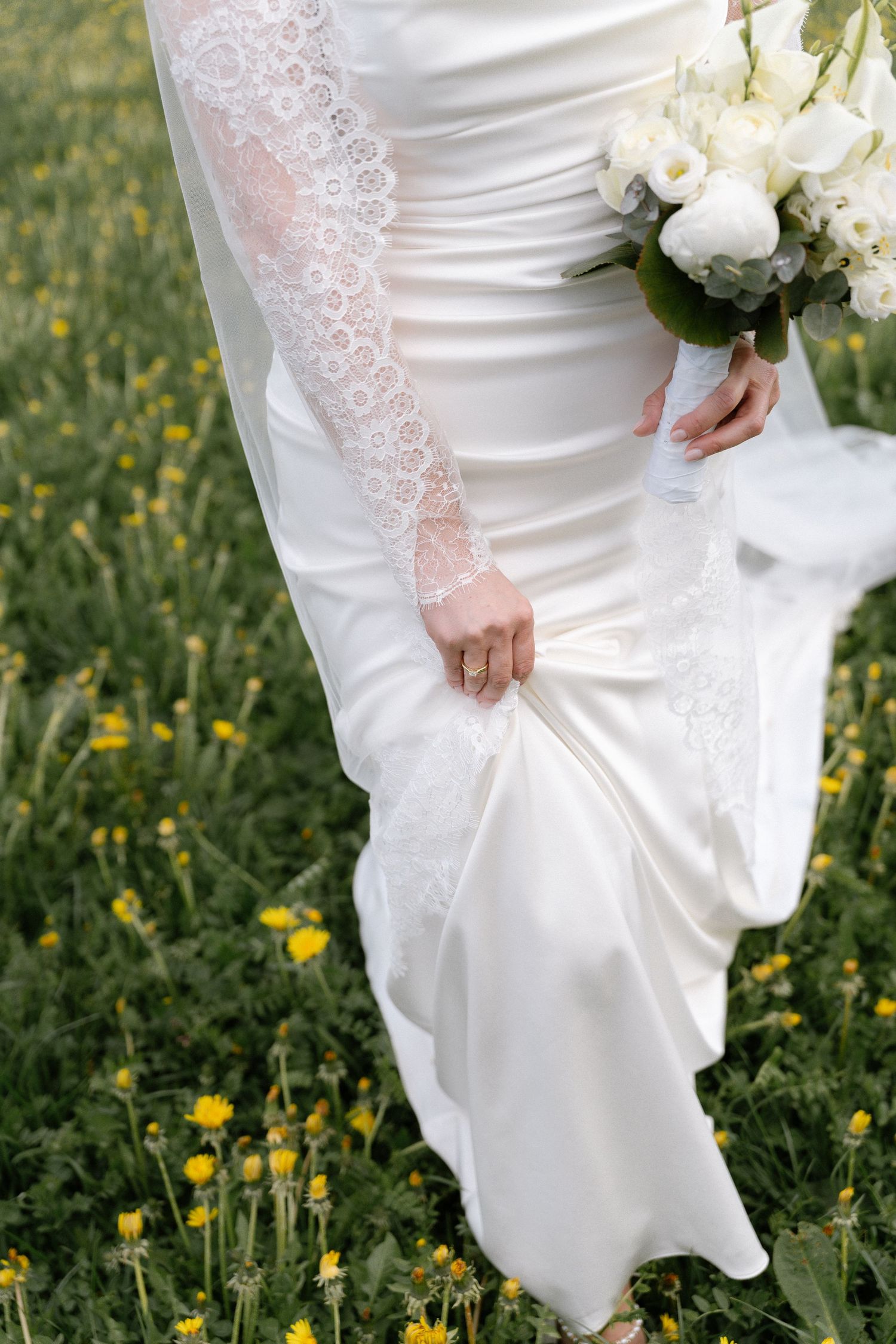 Bride in white lace wedding gown holding bouquet of white flowers while standing in green field with yellow wildflowers.