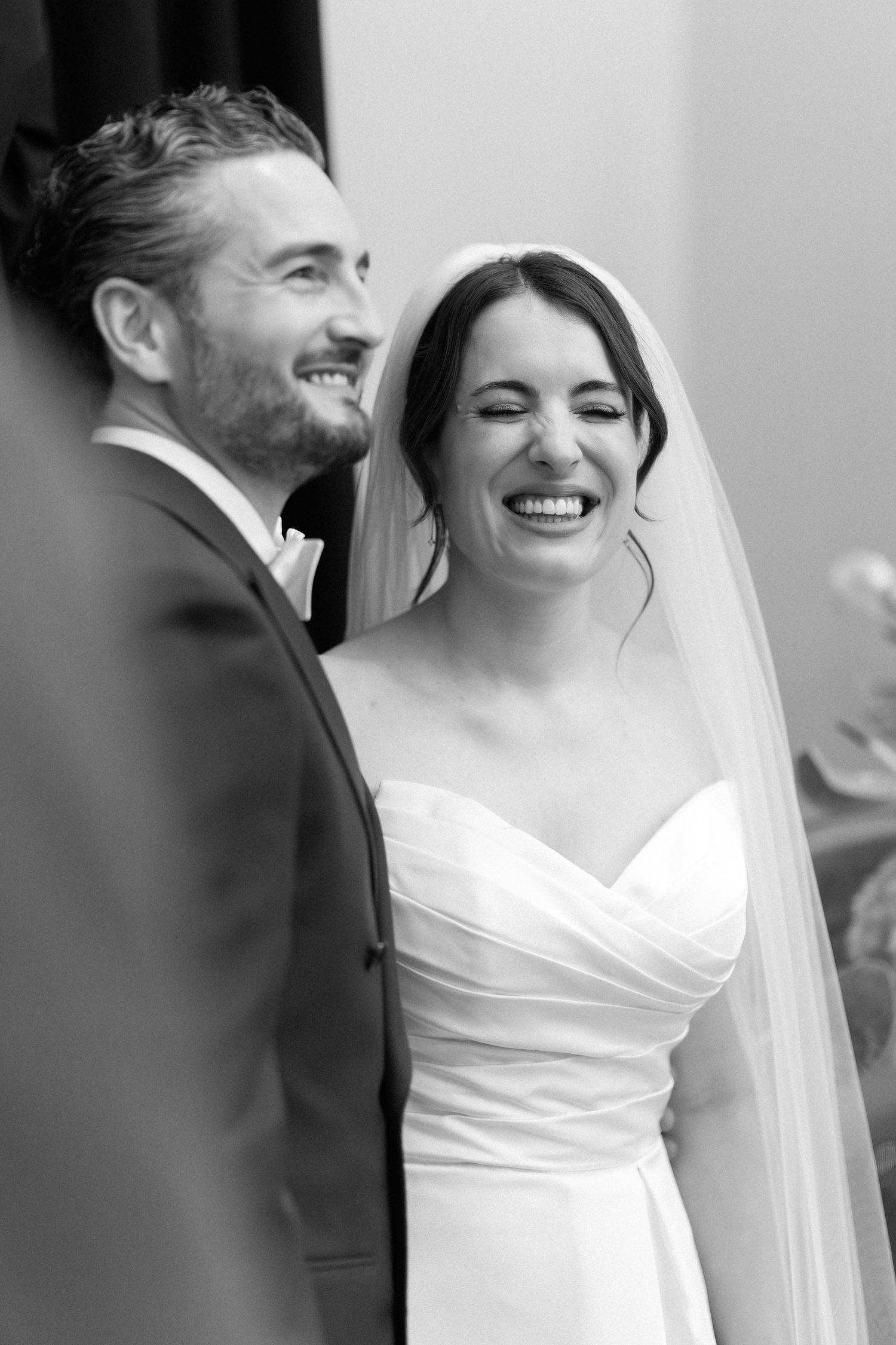 Black and white photo of a laughing bride in veil and groom in suit sharing a joyful moment during their wedding ceremony.