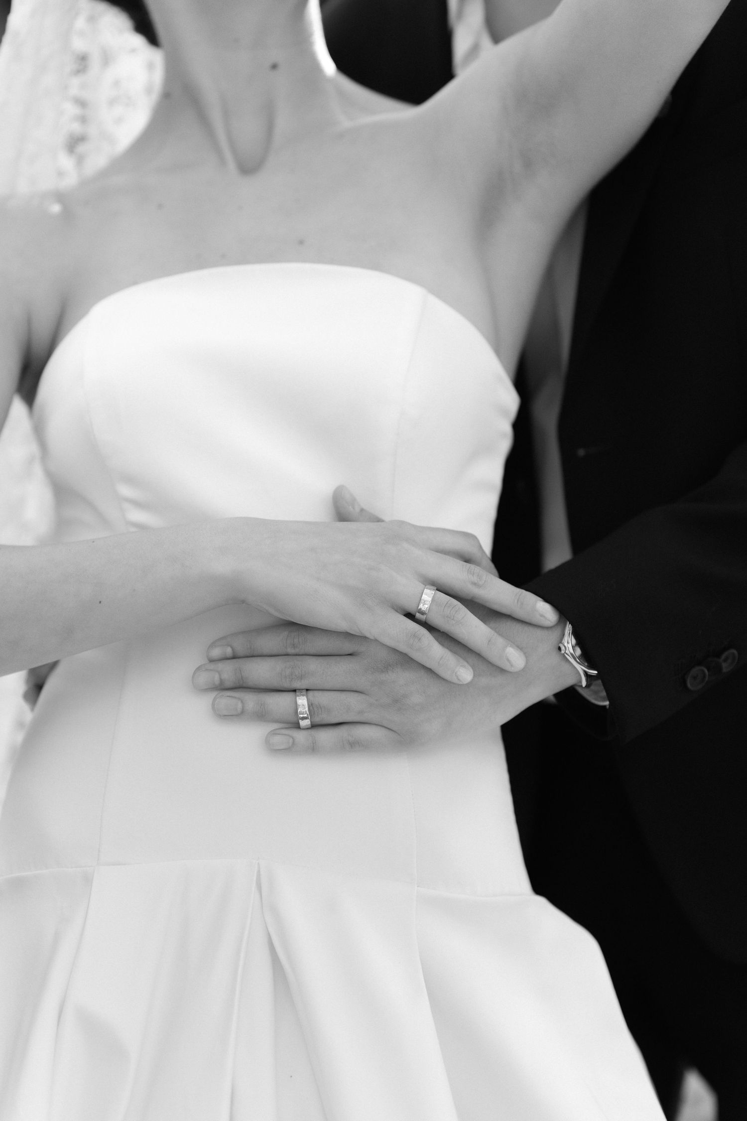 Black and white photo of newlyweds showing wedding rings, bride in strapless gown, groom in suit embracing her waist.