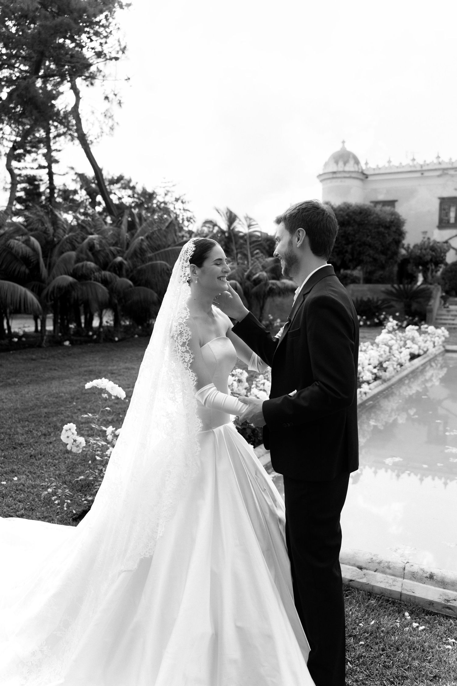 Black and white photo of a bride and groom sharing a tender moment in a lush garden with a reflecting pool.