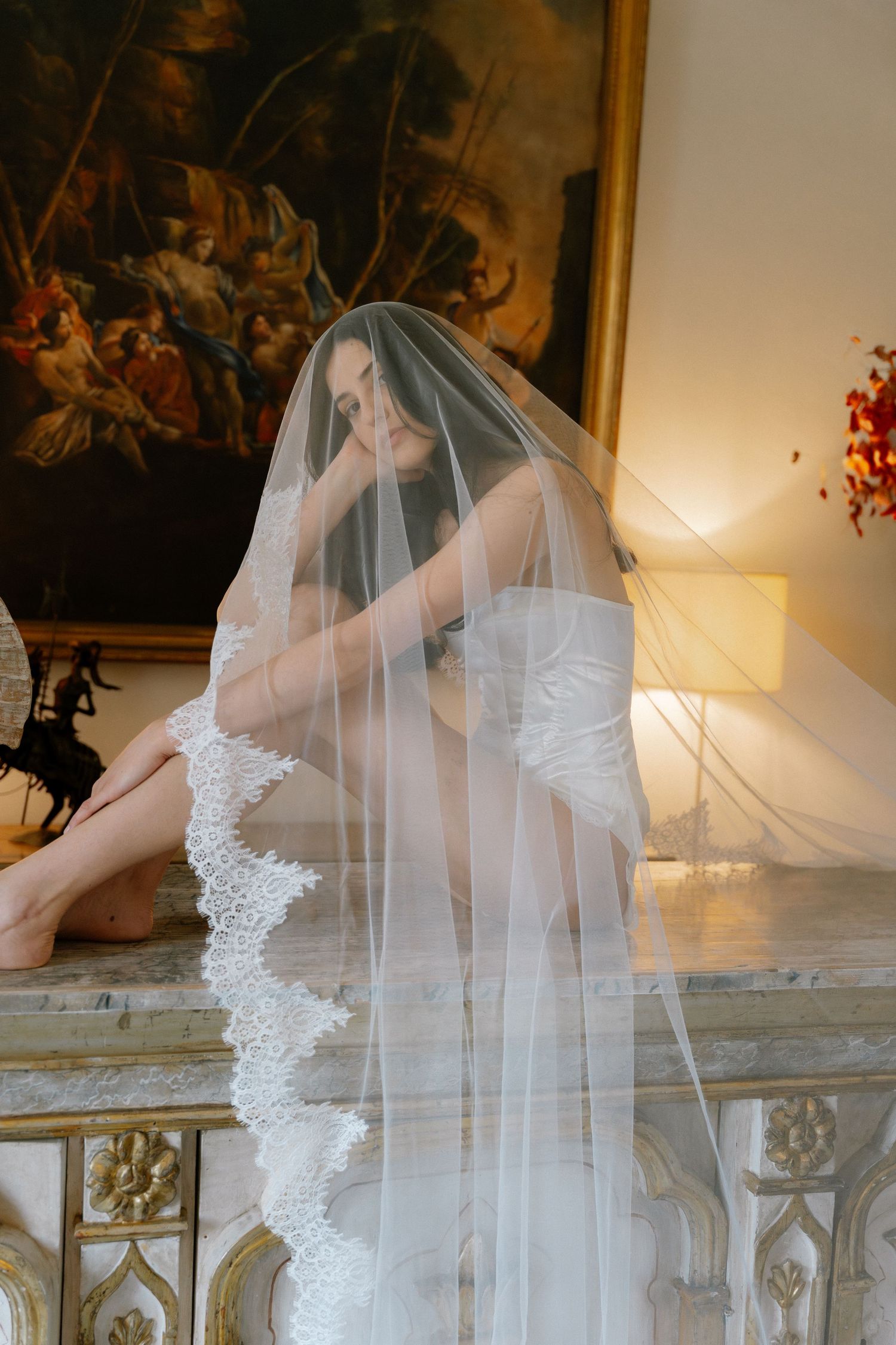 Bride in white lace wedding dress and veil seated on marble mantle before classic oil painting.