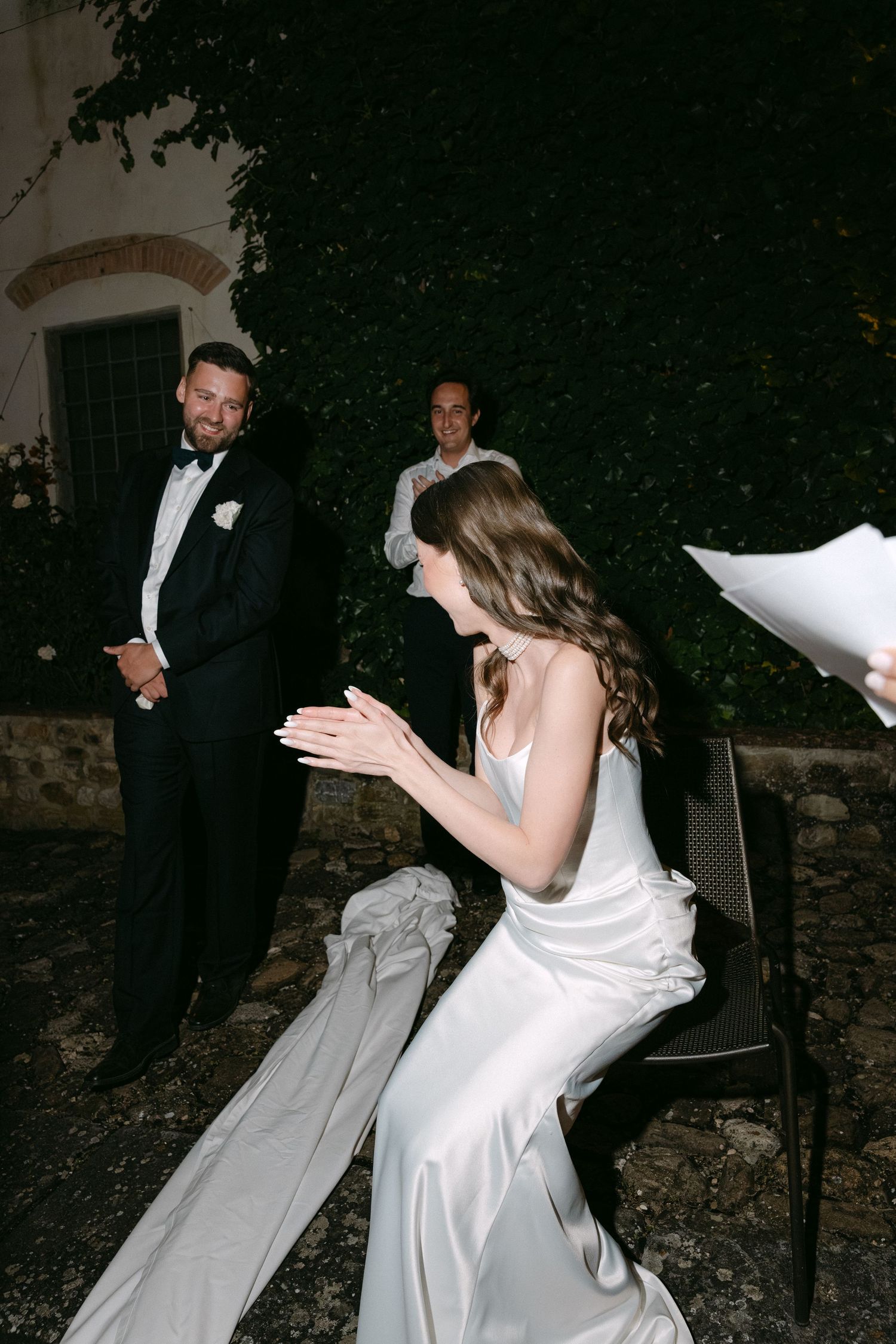 Bride in silk gown reads vows from paper while groom in tuxedo watches during outdoor evening wedding ceremony.