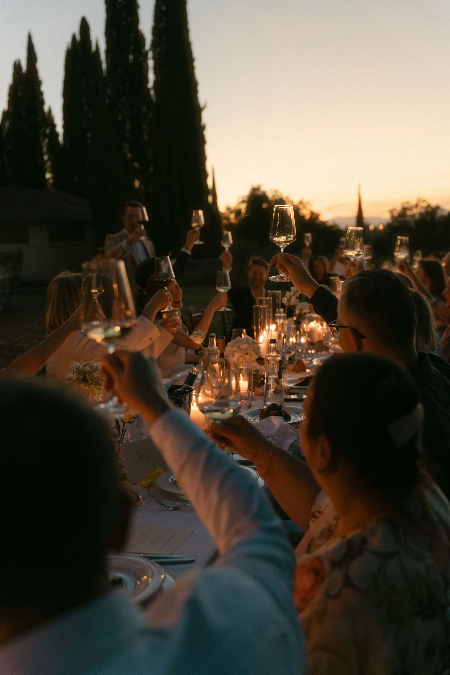 Guests raising wine glasses in a toast at an elegant outdoor dinner party at sunset with candlelit table and cypress trees.