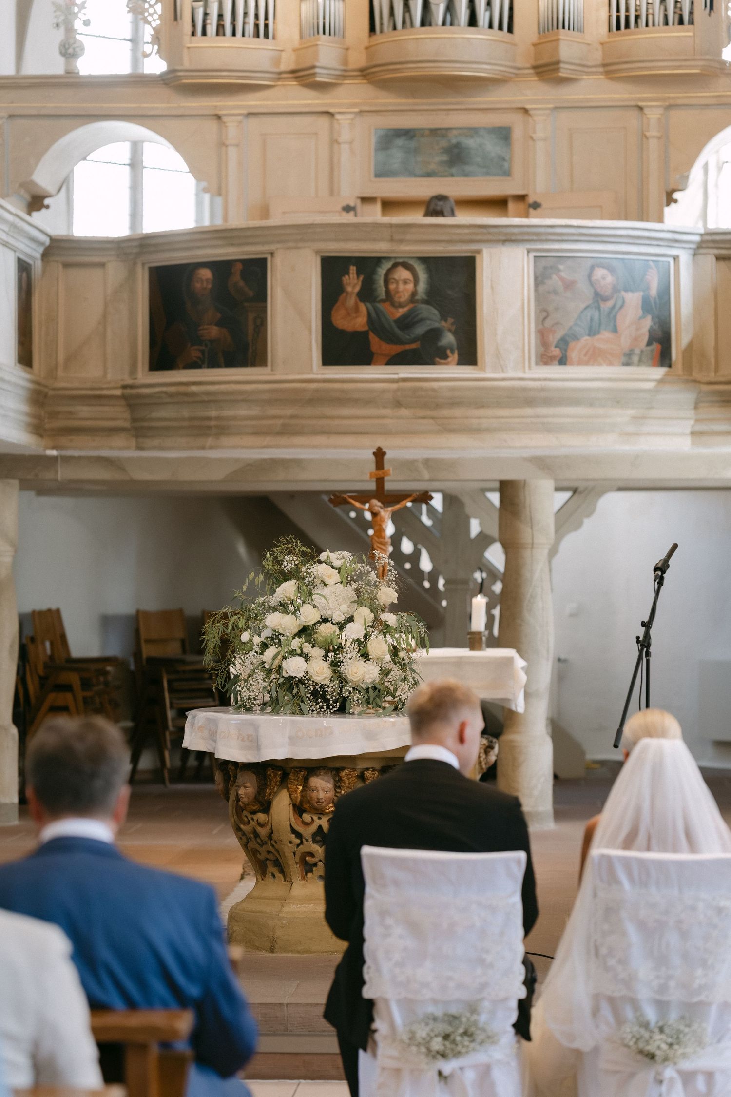 Bride in a veil and groom stand before a flower-draped altar with a crucifix during a wedding ceremony in a historic church.