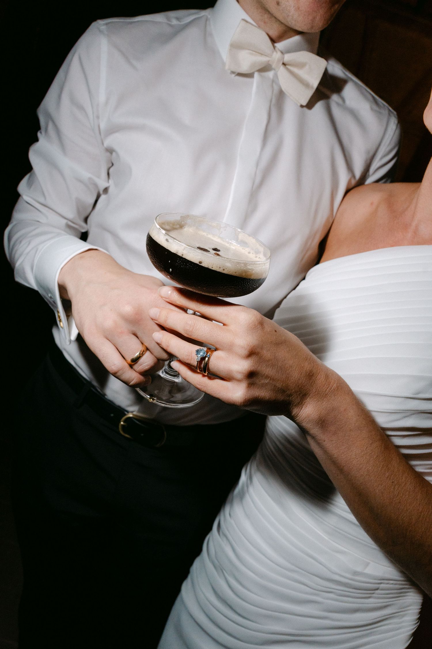 Couple in wedding attire holding an espresso martini, the groom in a white bow tie and the bride in a ruched white dress.
