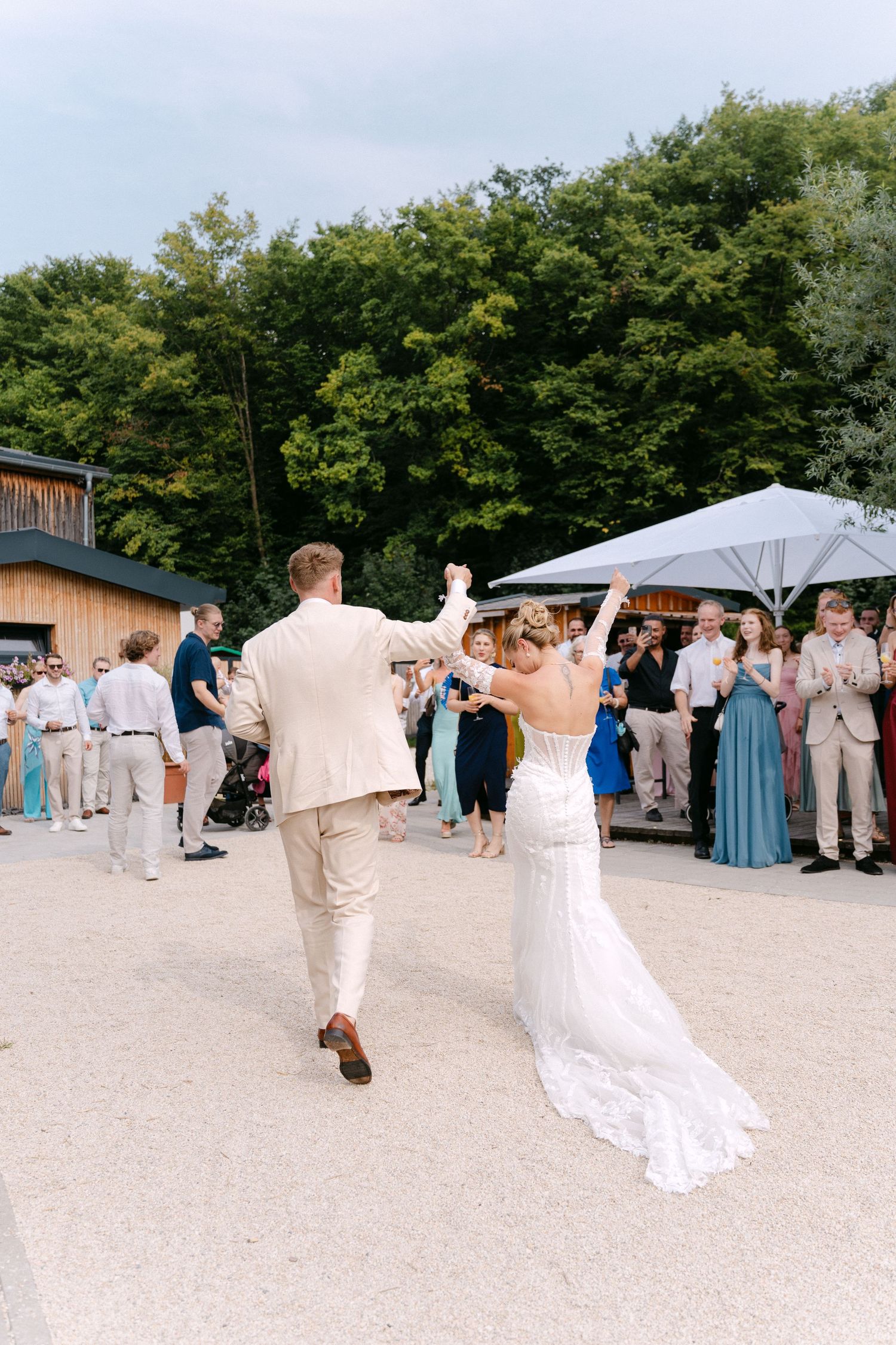 Wedding party dances together on sandy beach surrounded by trees during outdoor summer celebration.