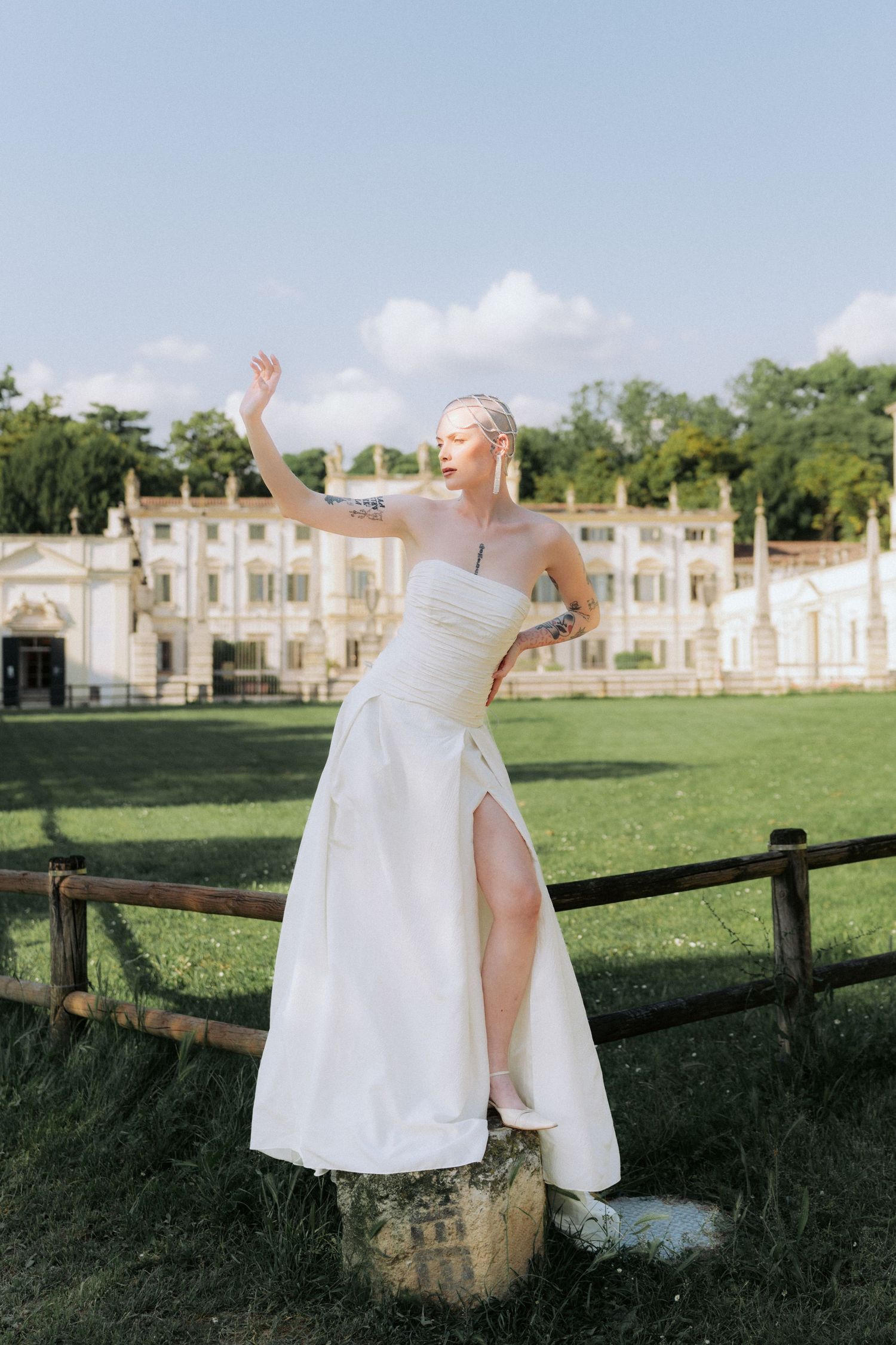 A figure in a white dress sits and poses on a rustic wooden fence in front of a stately building at sunset.