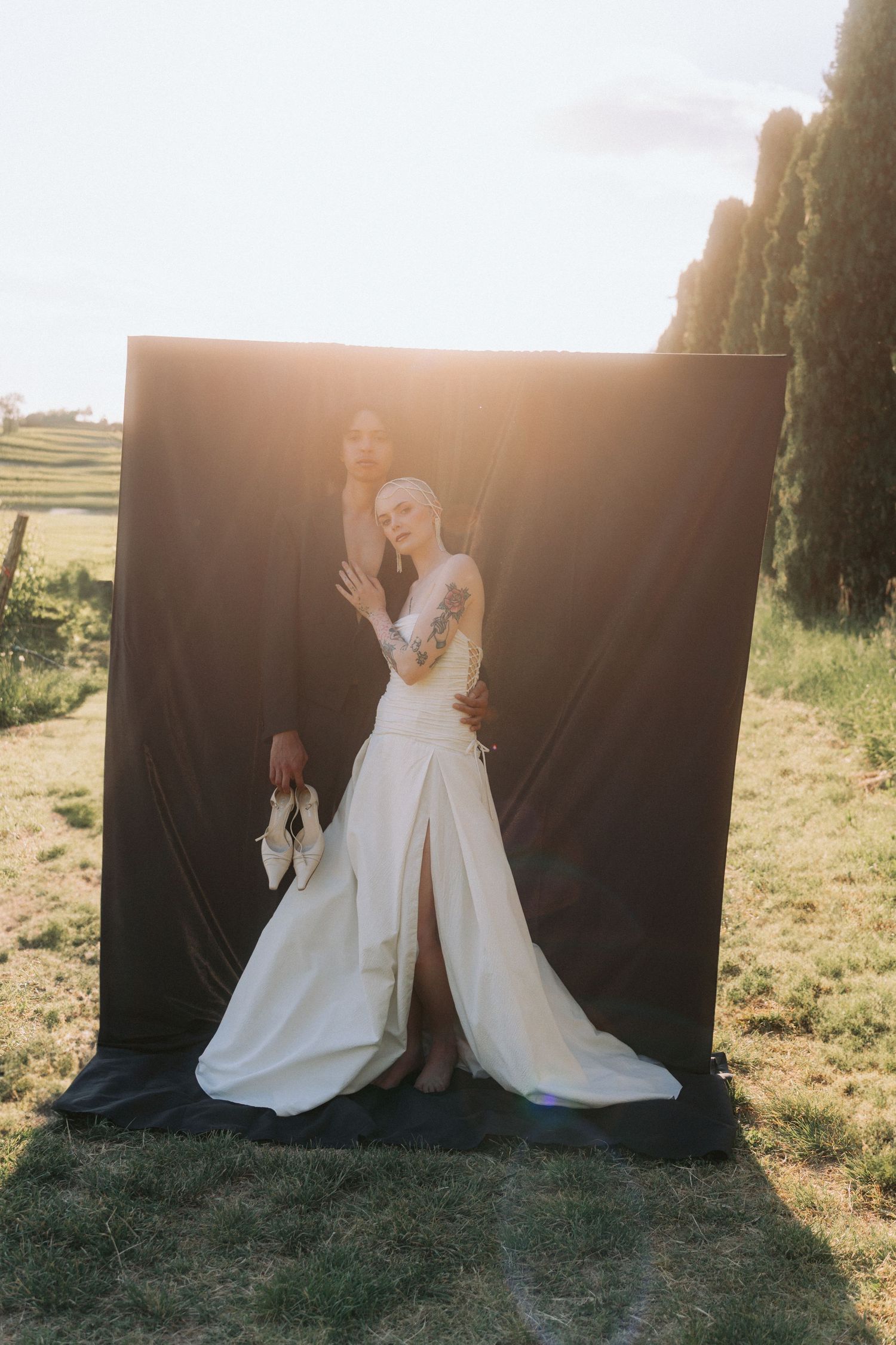 Two people in flowing white wedding dresses sit together on grass at sunset with dramatic backlighting.