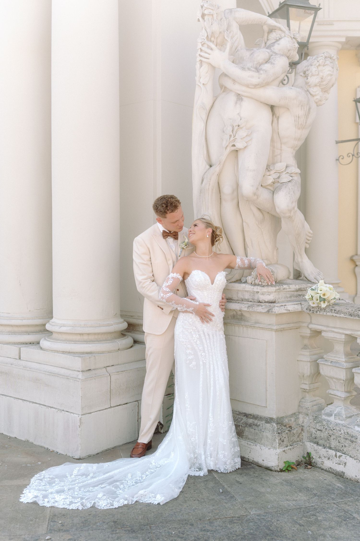 Elegant wedding couple poses by ornate classical stone statue and white columns in light beige formal attire. Schloss Ludwigsburg