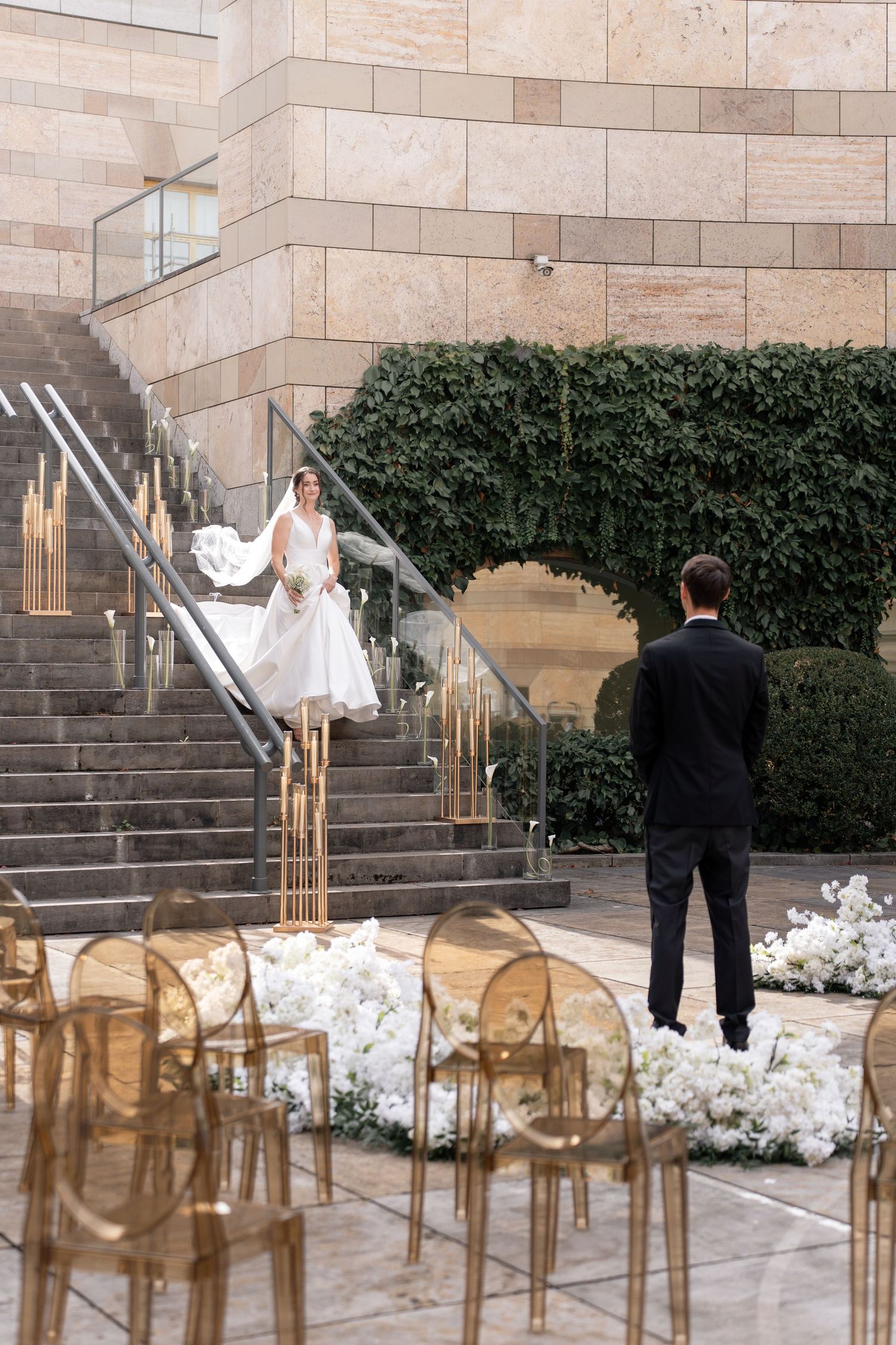 An elegant outdoor wedding setup with ghost chairs and white floral arrangements on stone steps with a modern building backdrop.