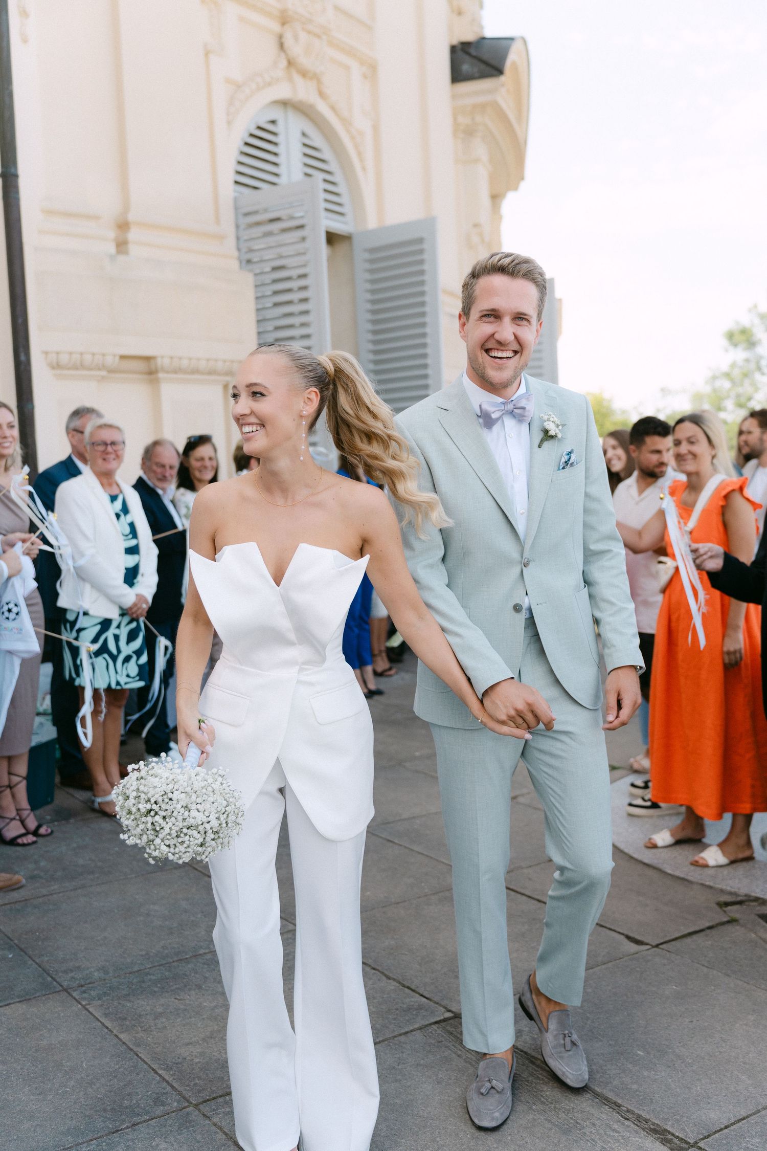 A happy couple exits their wedding ceremony, with the bride in a white jumpsuit and groom in light blue suit. Schloss Solitude