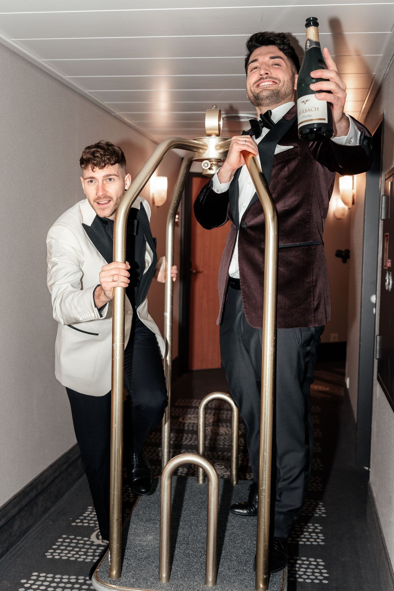 Two men in formal attire pose playfully with wine bottles in a narrow hallway with metal railings.