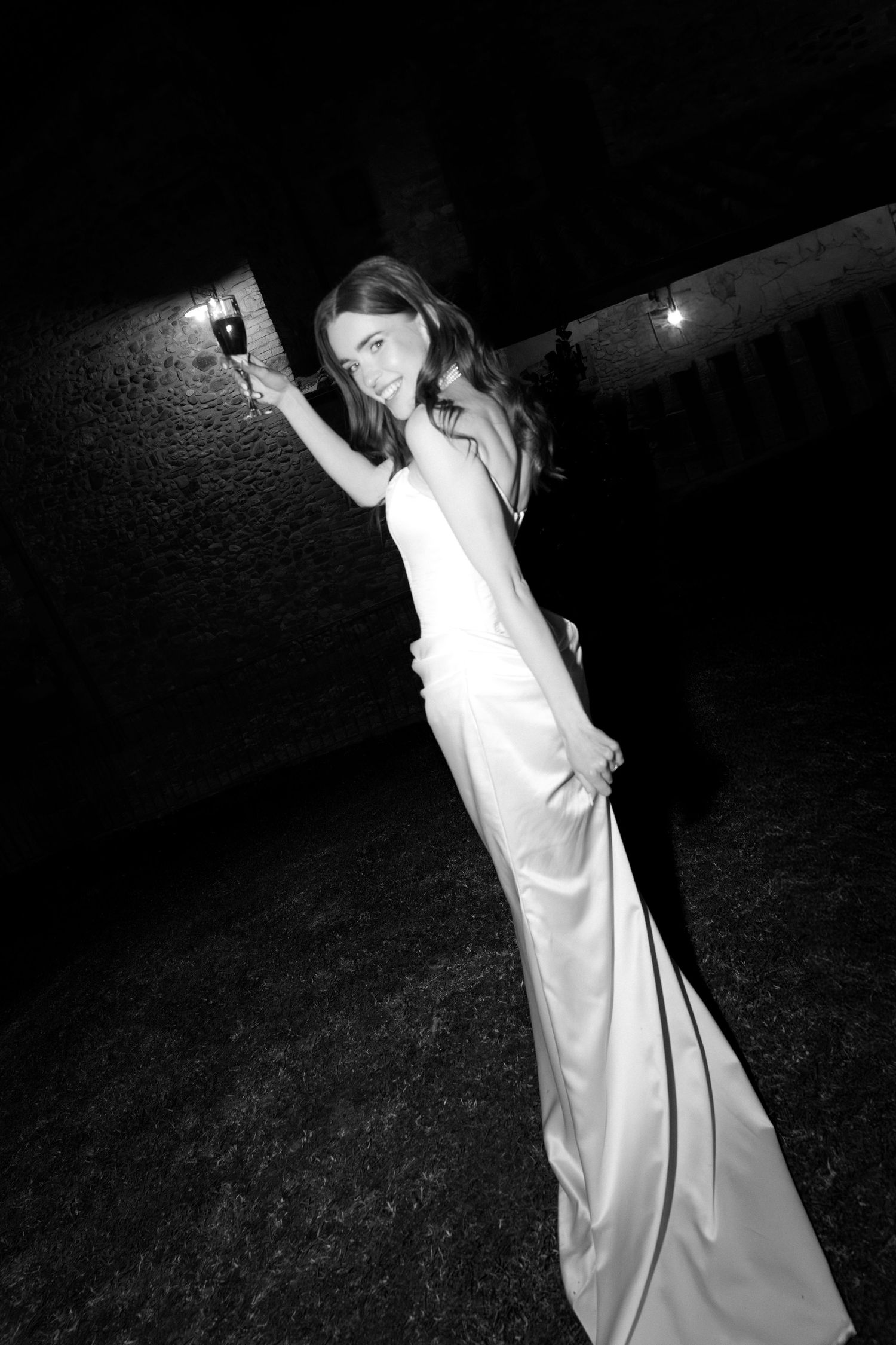 Woman in elegant white gown raising a wine glass outdoors at night, black and white photography.