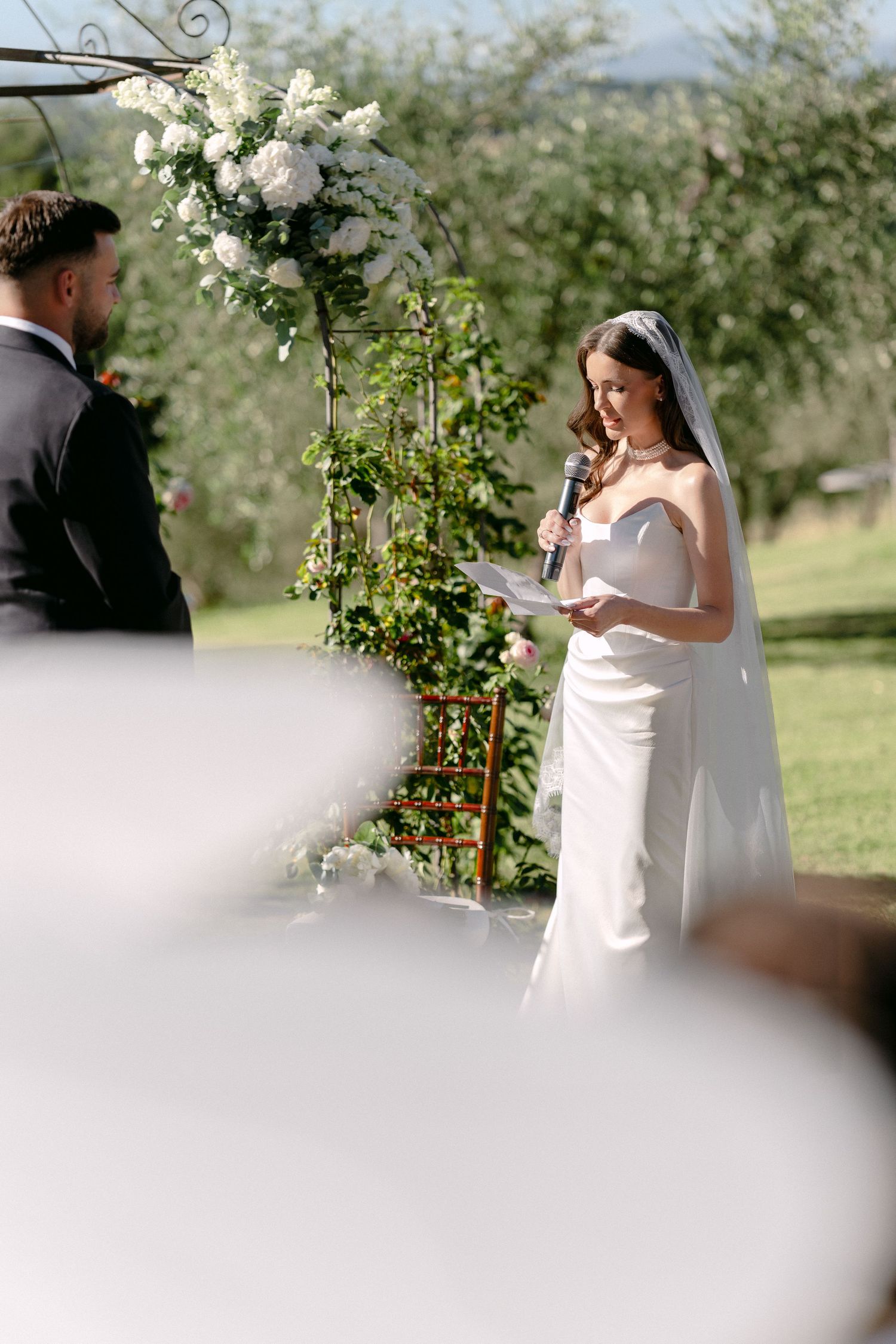 Bride reading vows at outdoor wedding ceremony with floral arch backdrop and lush greenery.