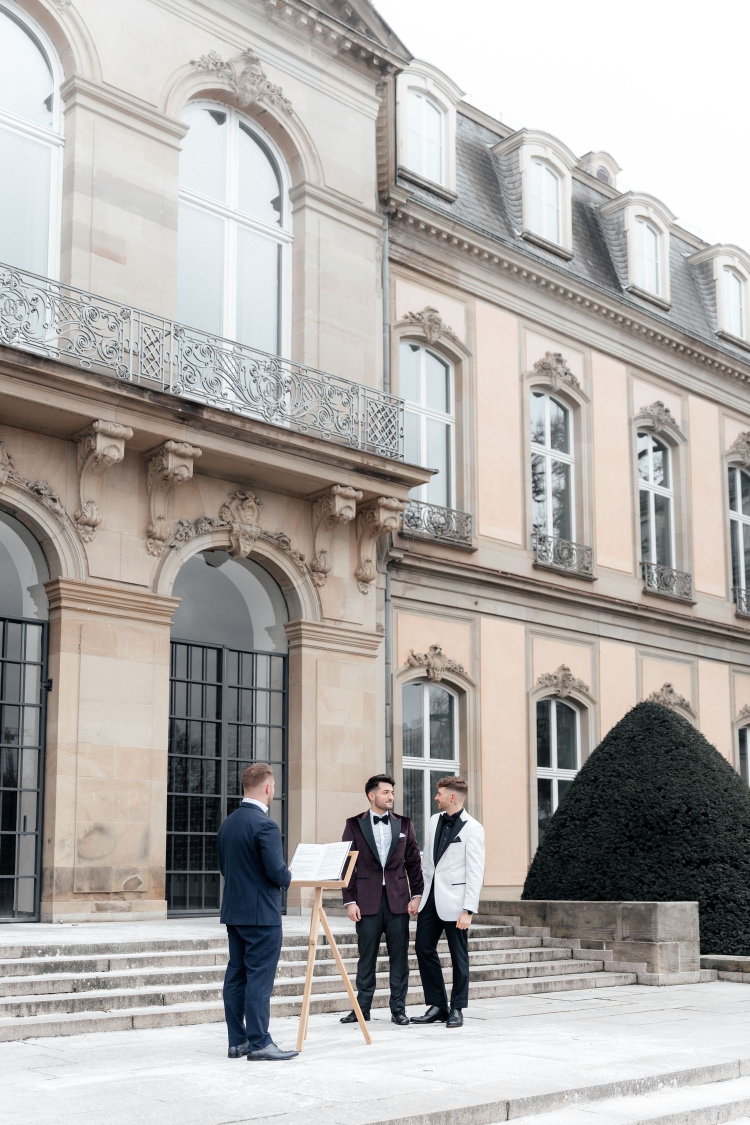 Three people in formal attire standing on stone steps of an elegant French-style mansion with ornate architectural details.