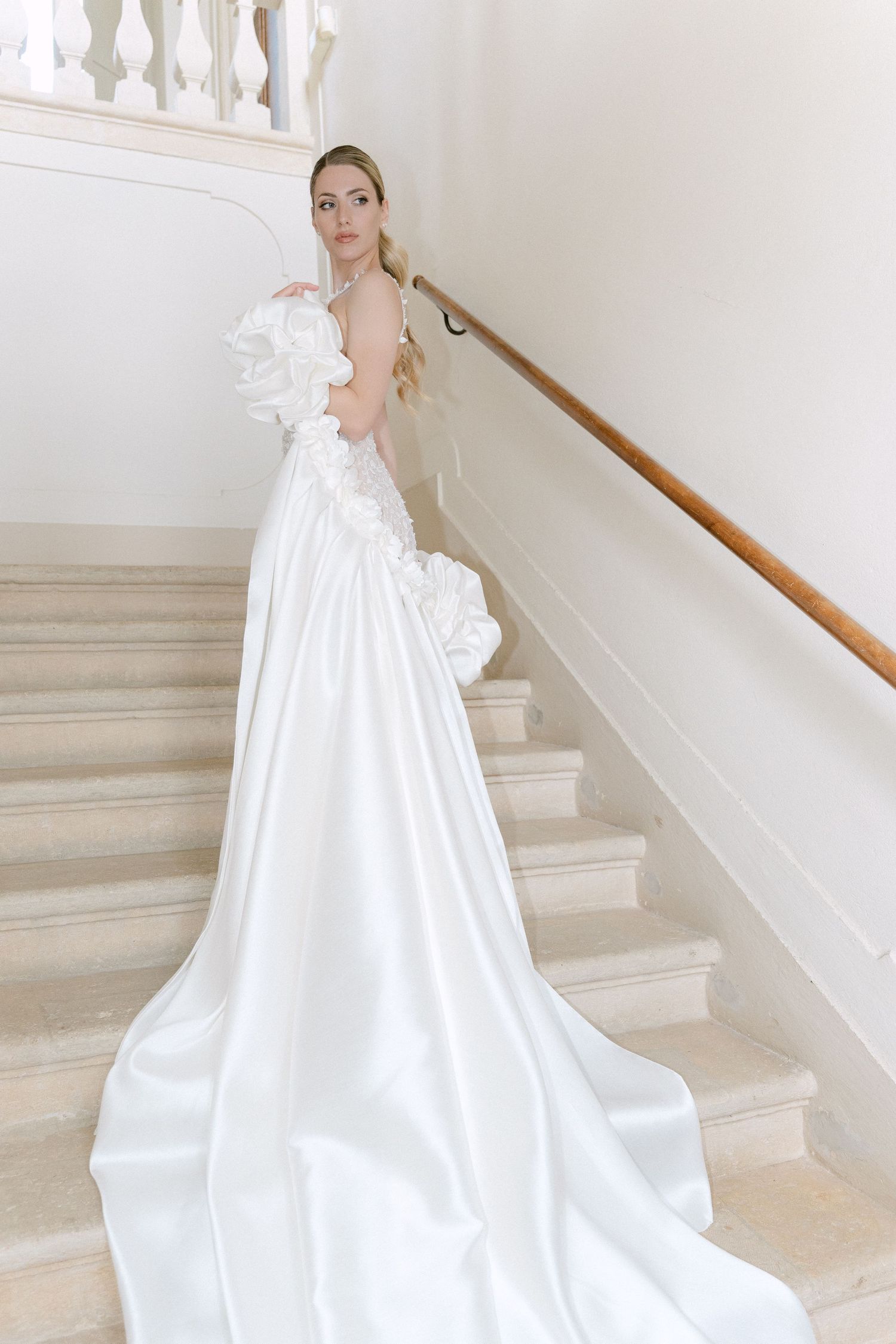 A bride in a flowing white wedding gown with floral details poses elegantly on a grand marble staircase. Wedding Verona