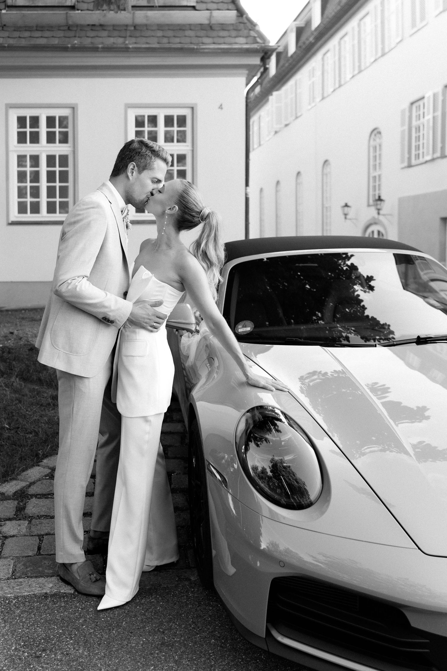 A couple in white formal attire poses romantically with a sleek white Porsche sports car on their wedding day. Schloss Solitude