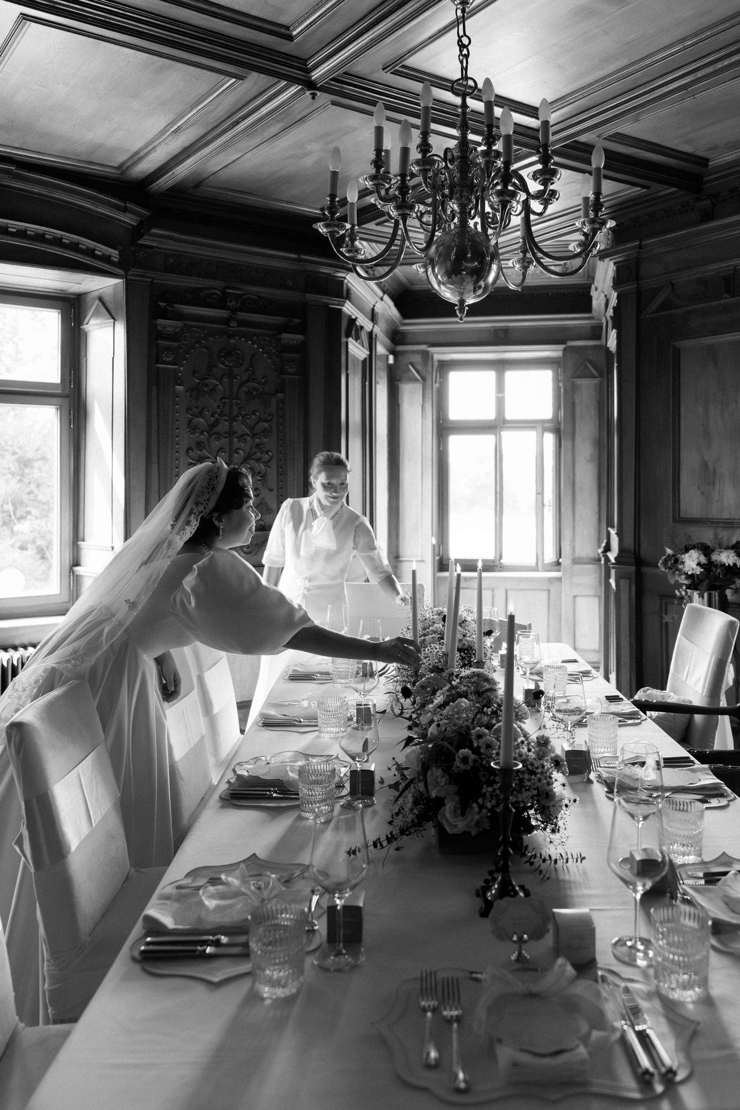 Black and white photo of a bride arranging a long elegantly set wedding reception table in a grand wood-paneled dining room.