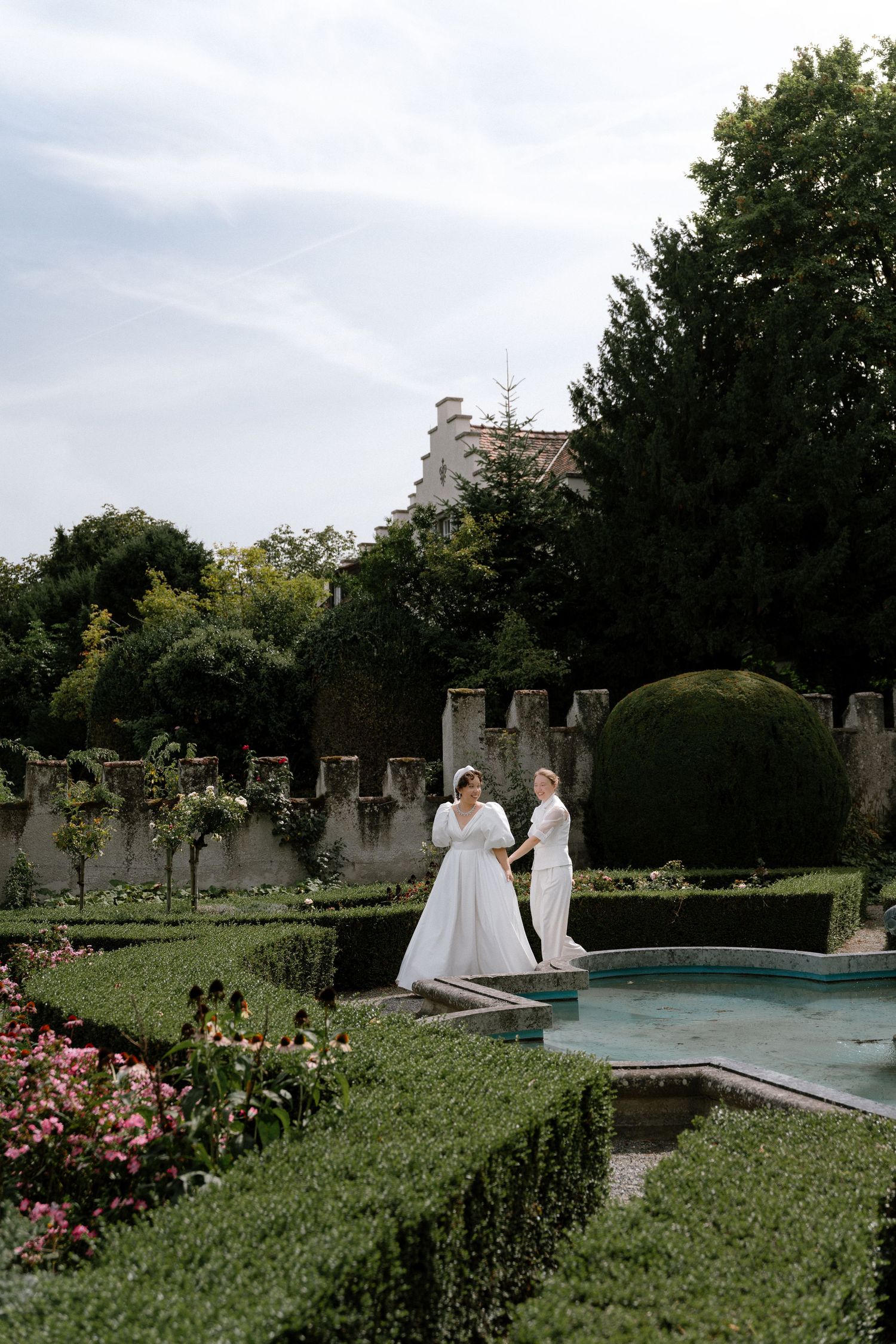 Two brides in white gowns hold hands in a formal garden with manicured hedges, fountain, and historic manor backdrop.