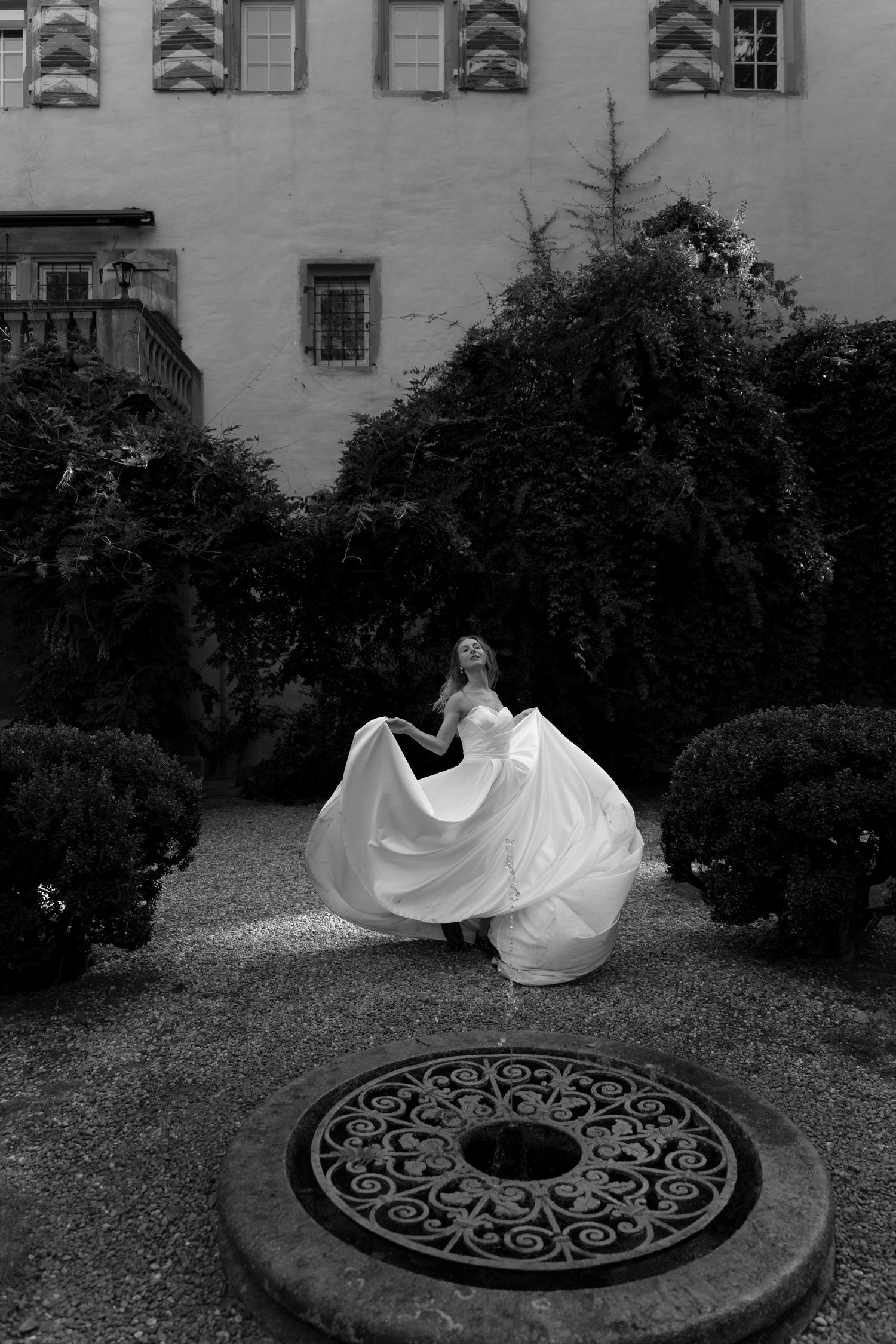 Black and white photo of a bride in a flowing gown twirling in a courtyard garden with ivy, shrubs and an ornate iron well cover.