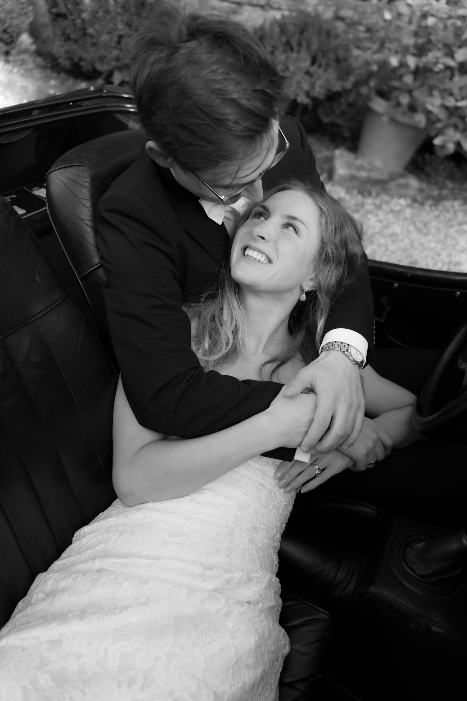 Black and white photo of a bride in a lace wedding dress smiling up at a man in a suit embracing her in a vintage convertible.