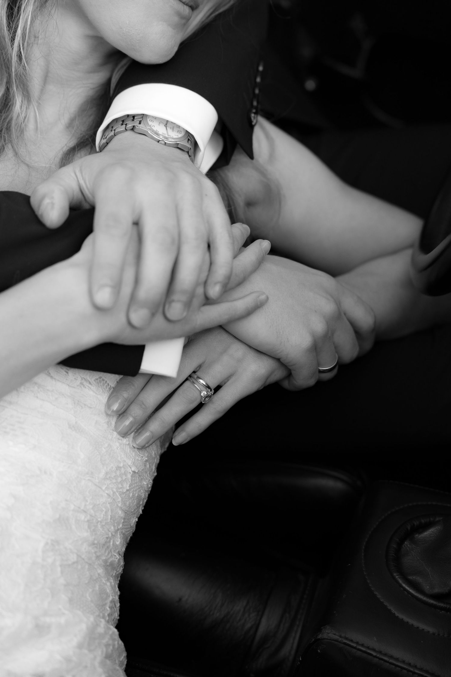 Black and white close-up of a bride and groom's clasped hands with a wedding ring and watch, resting on a leather seat.