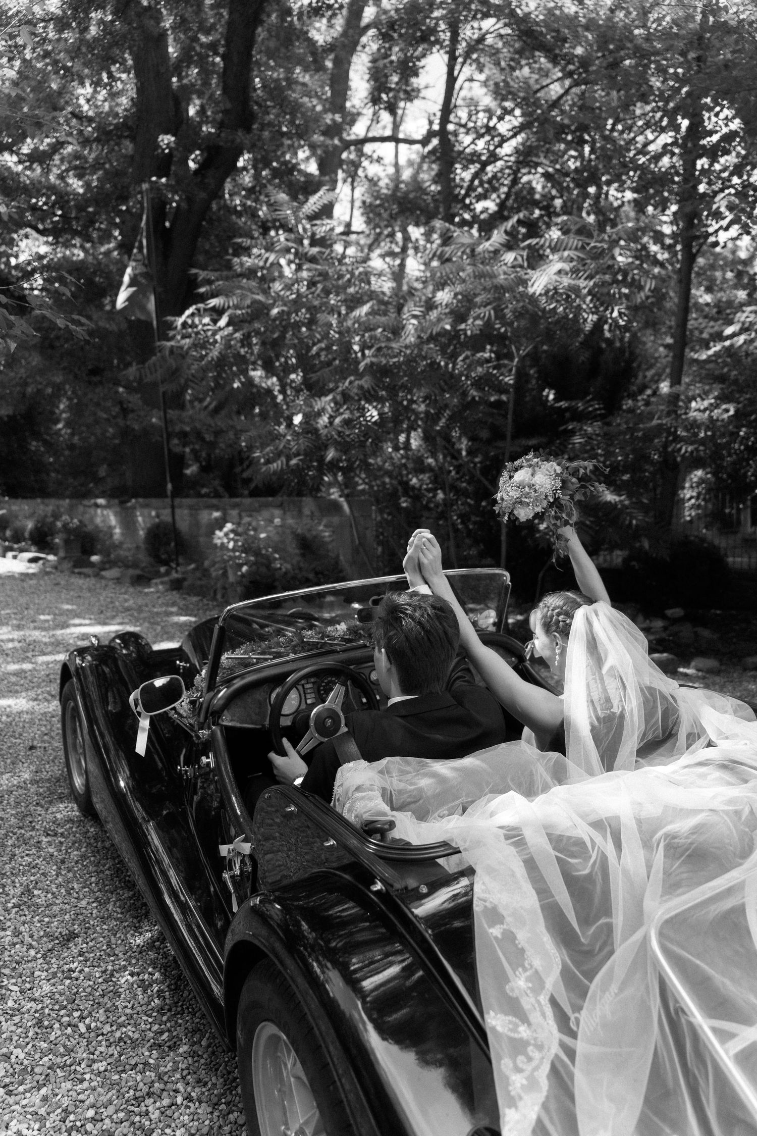 Black and white photo of a bride with her flowing veil raised bouquet in a vintage convertible car at an outdoor wedding.