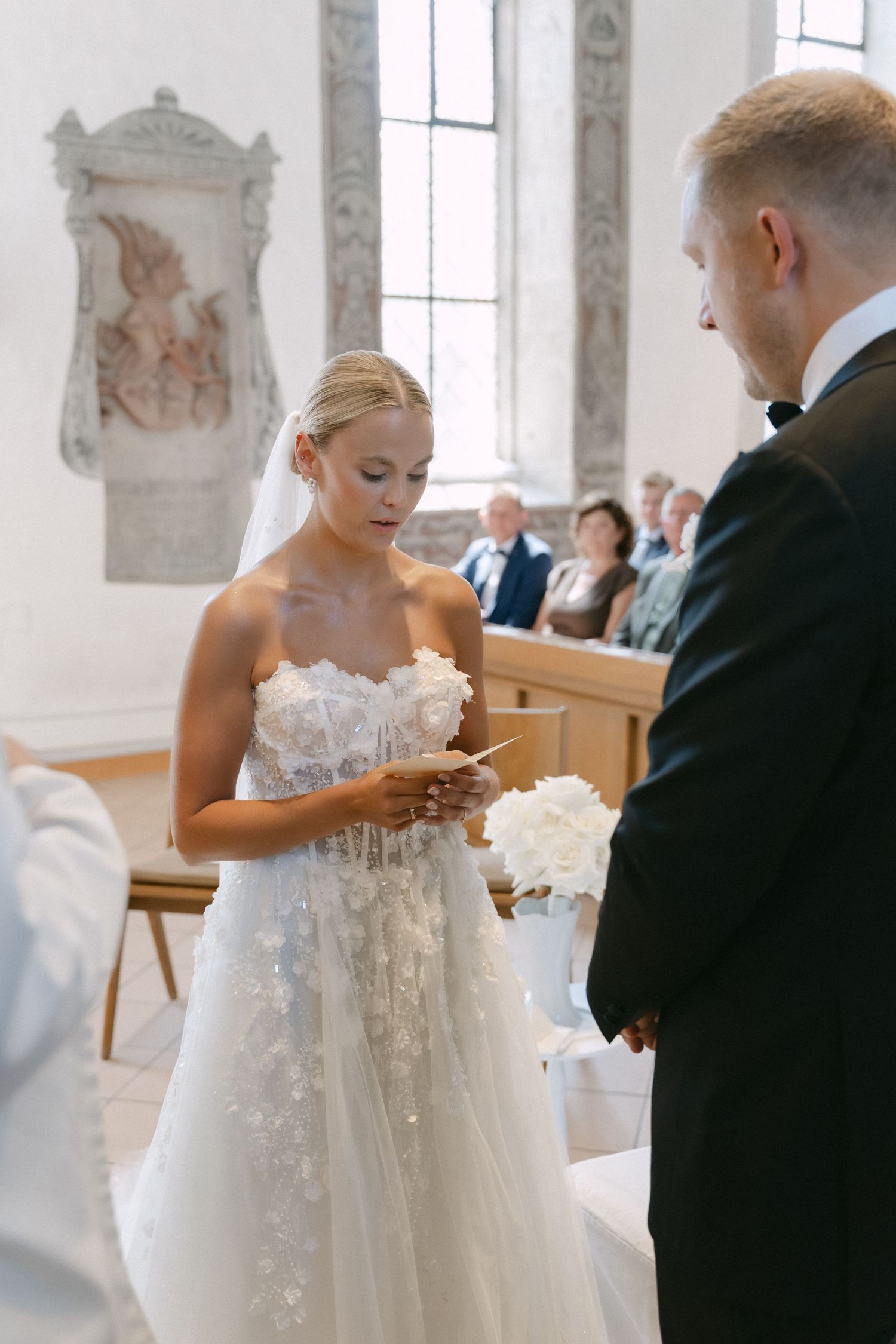 Bride in a strapless floral lace wedding gown reads her vows to the groom in a black suit during a ceremony in a bright stone church.