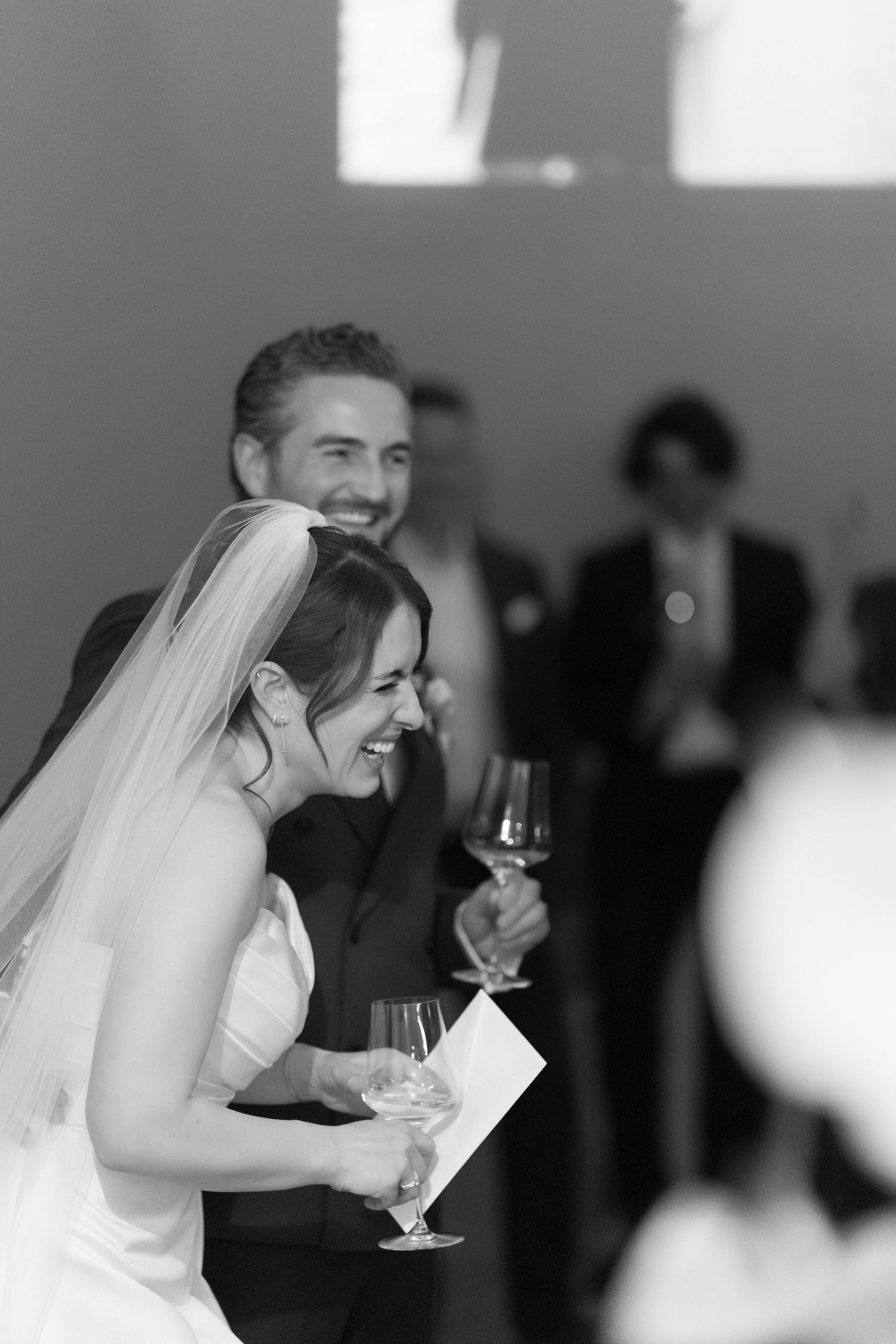 Black and white photo of a laughing bride holding champagne and papers at her wedding reception.