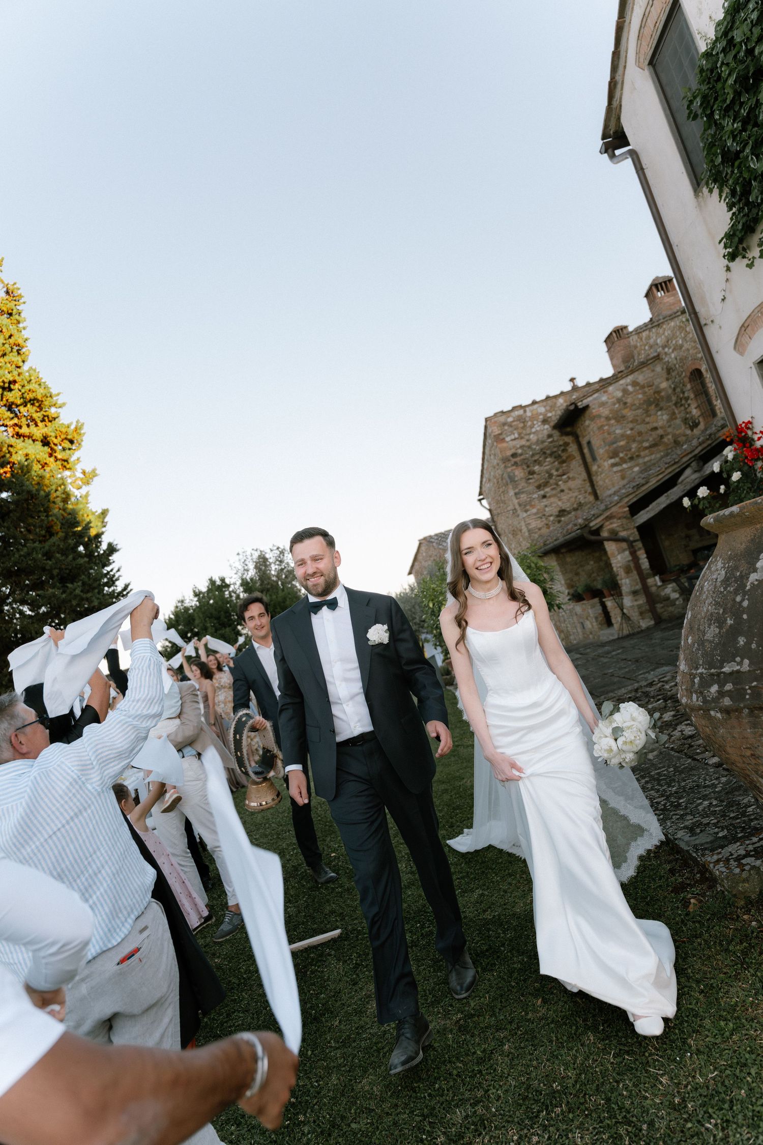 Bride and groom walk through cheering guests waving white napkins at outdoor wedding reception near rustic stone building.