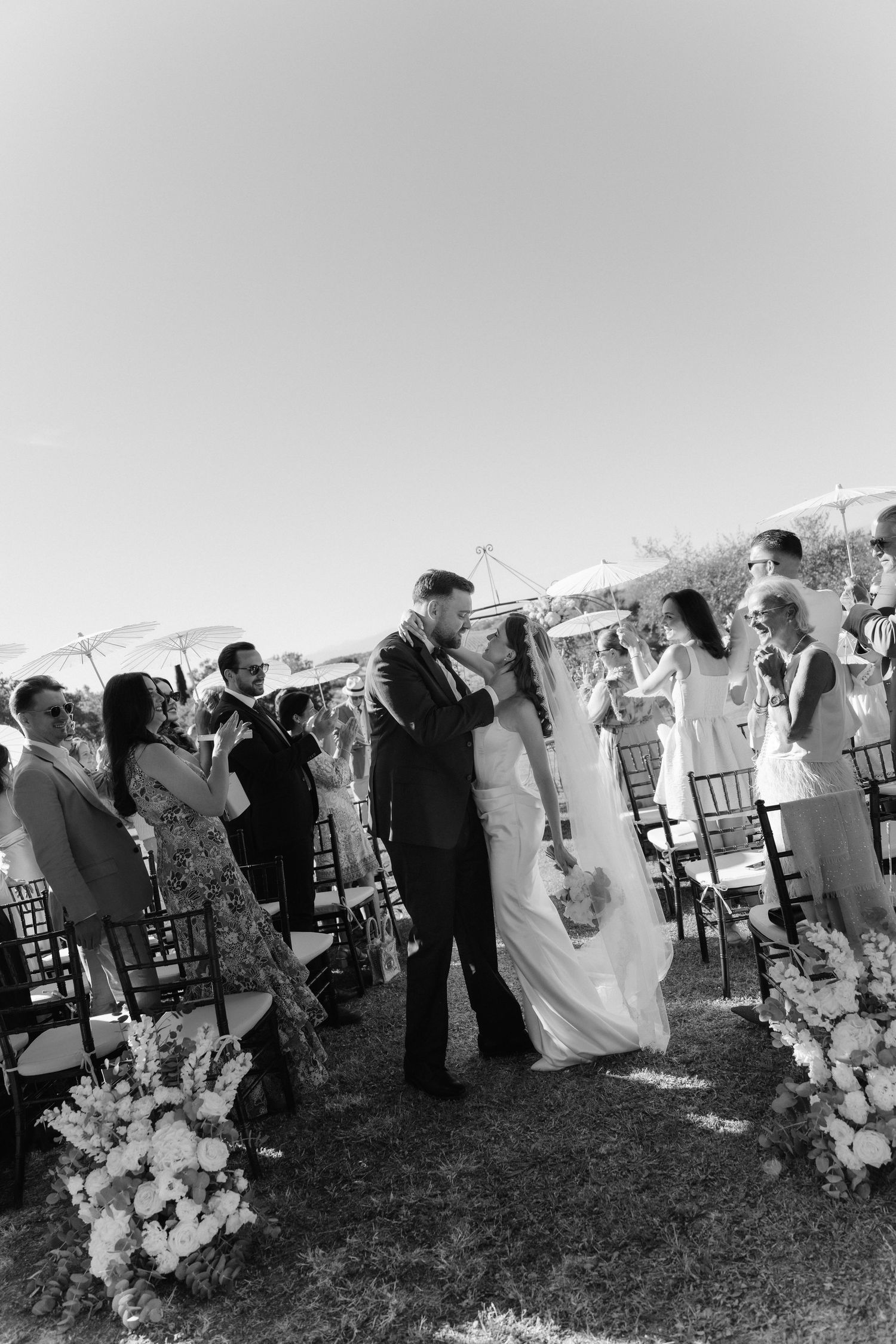 Black and white outdoor wedding ceremony photo of bride and groom sharing first kiss while guests celebrate.