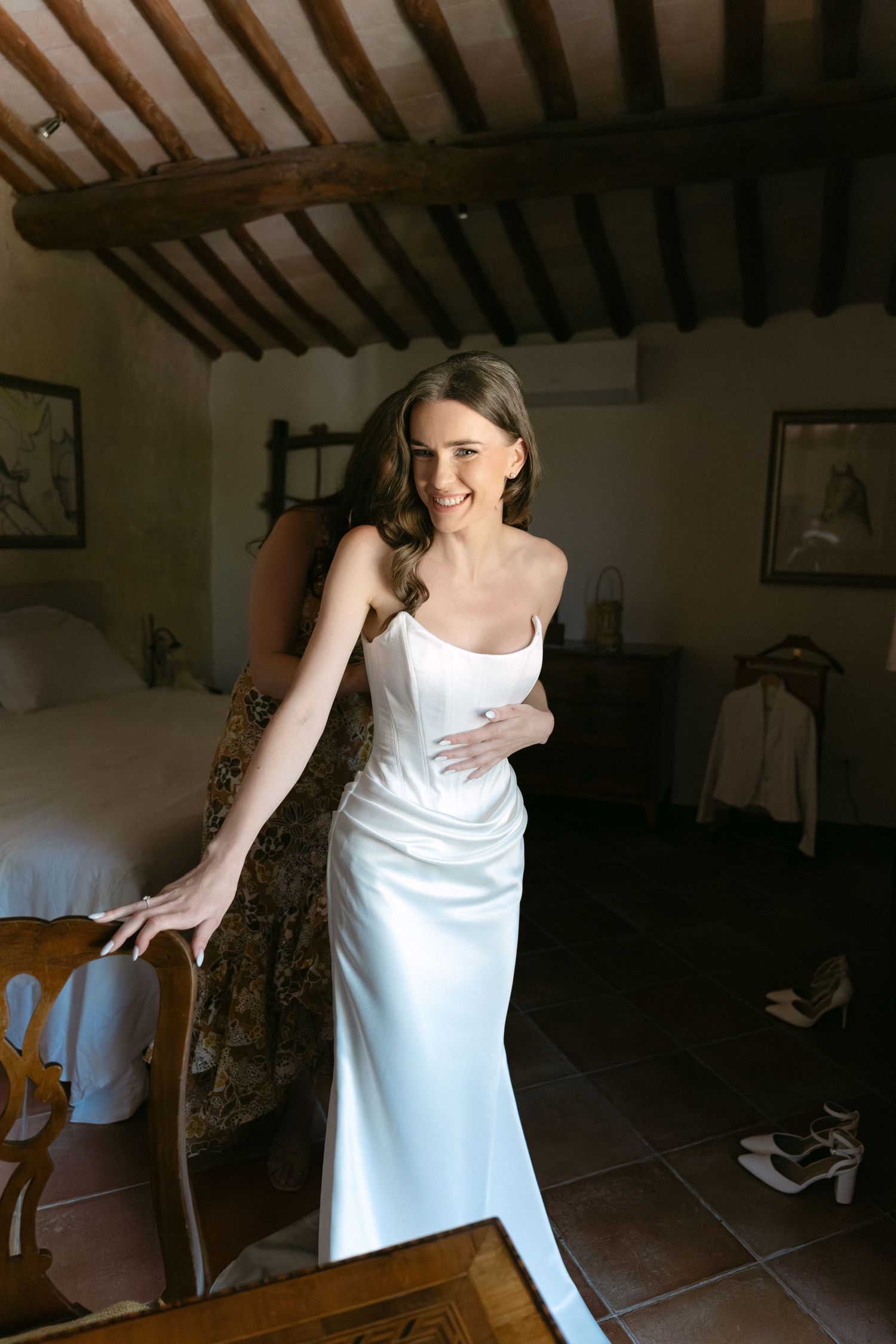 Smiling bride in elegant white satin slip wedding dress getting ready in rustic Tuscan villa bedroom.