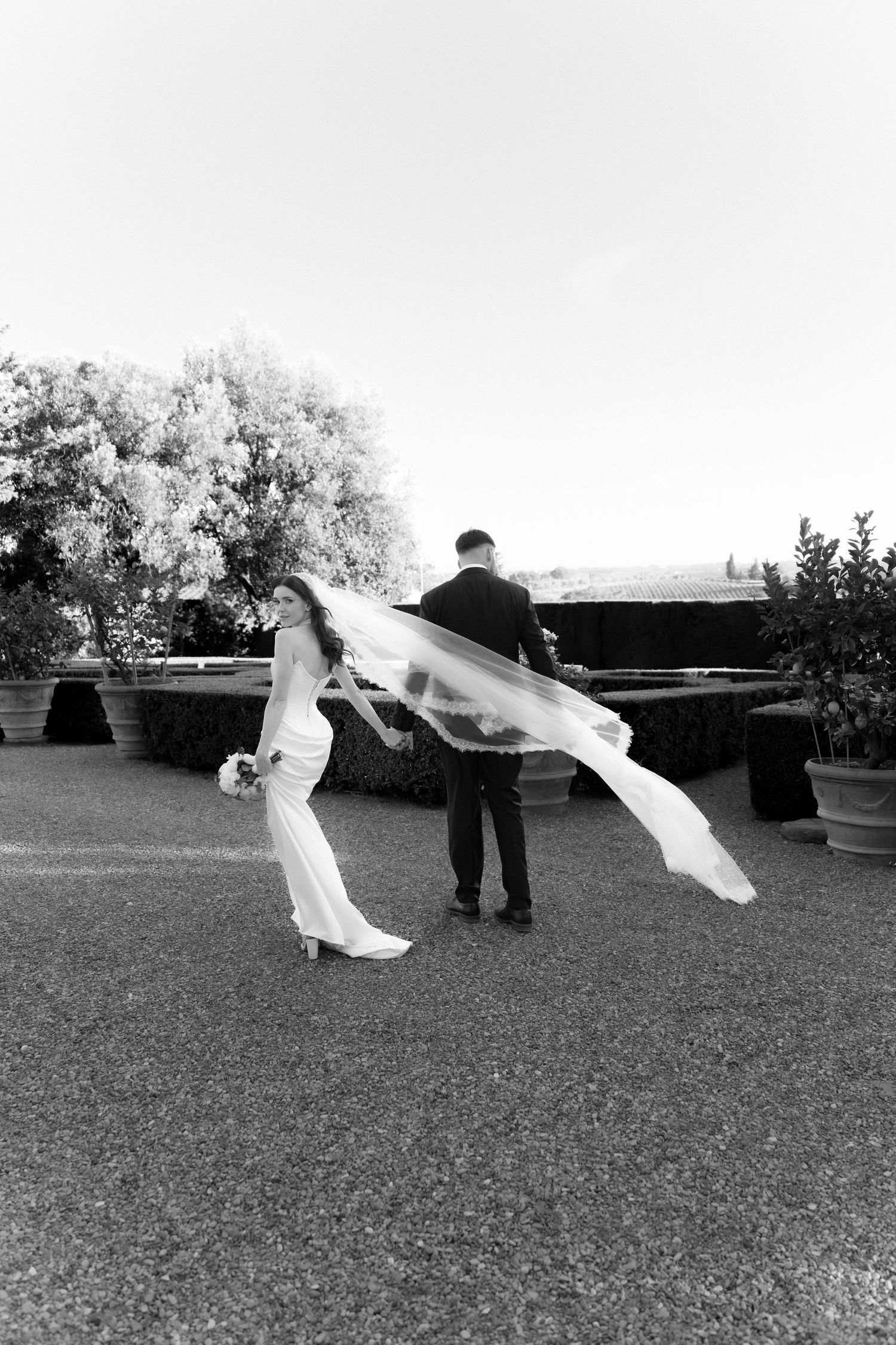 Black and white photo of bride and groom outdoors, veil flowing in wind, lush garden backdrop.