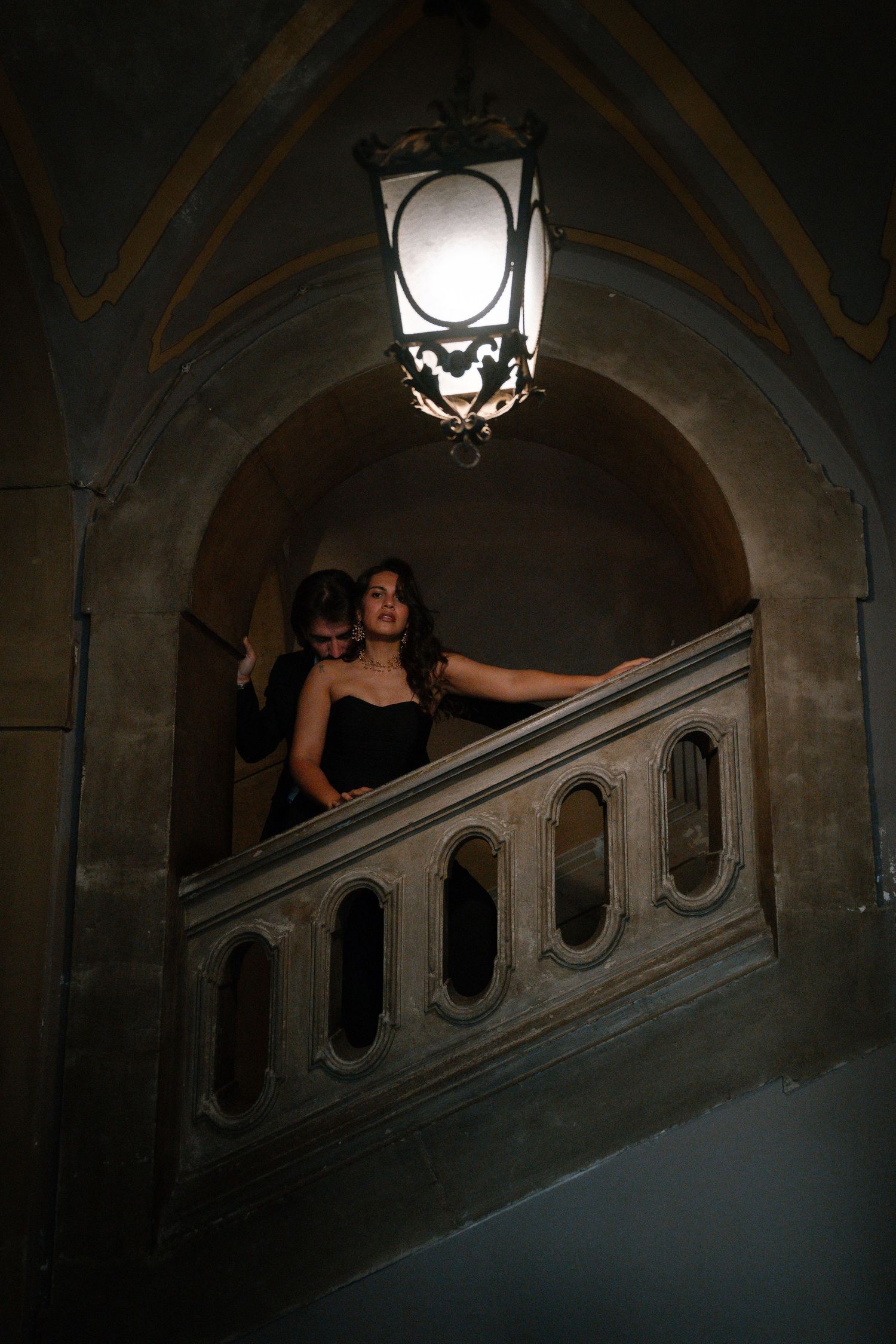 Couple in elegant black attire standing on ornate stone staircase beneath vintage lantern in dimly lit archway.