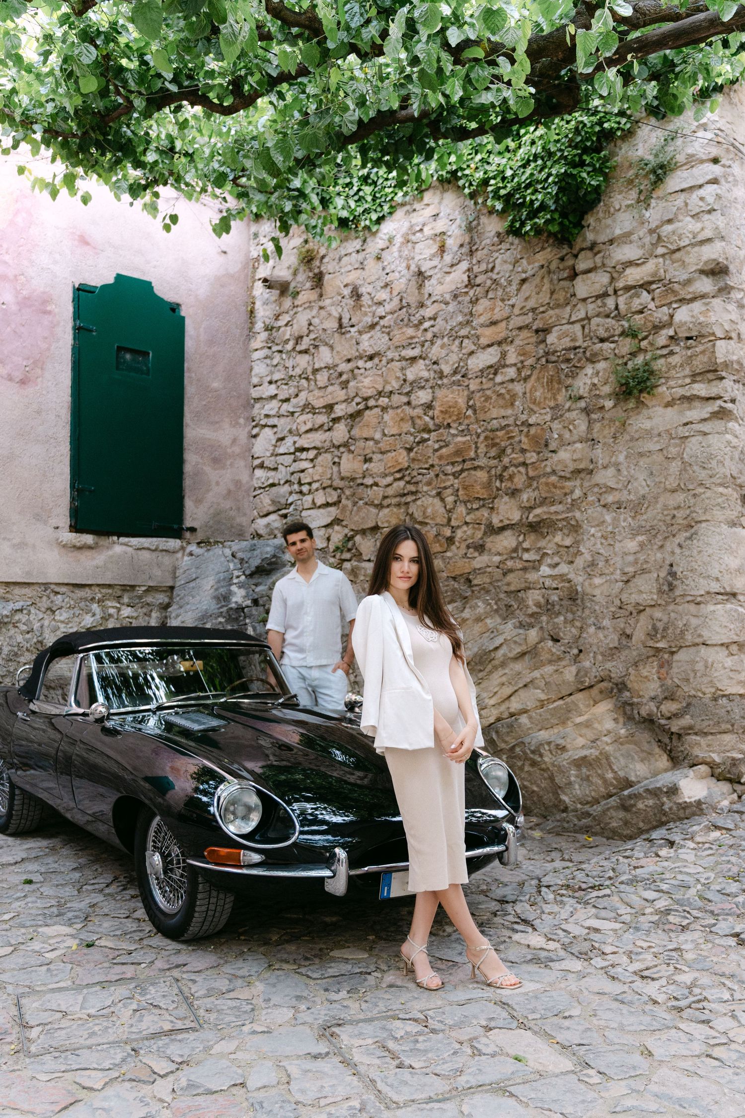 A classic black Jaguar E-Type parked against a rustic stone wall with green foliage and a teal door in the background.