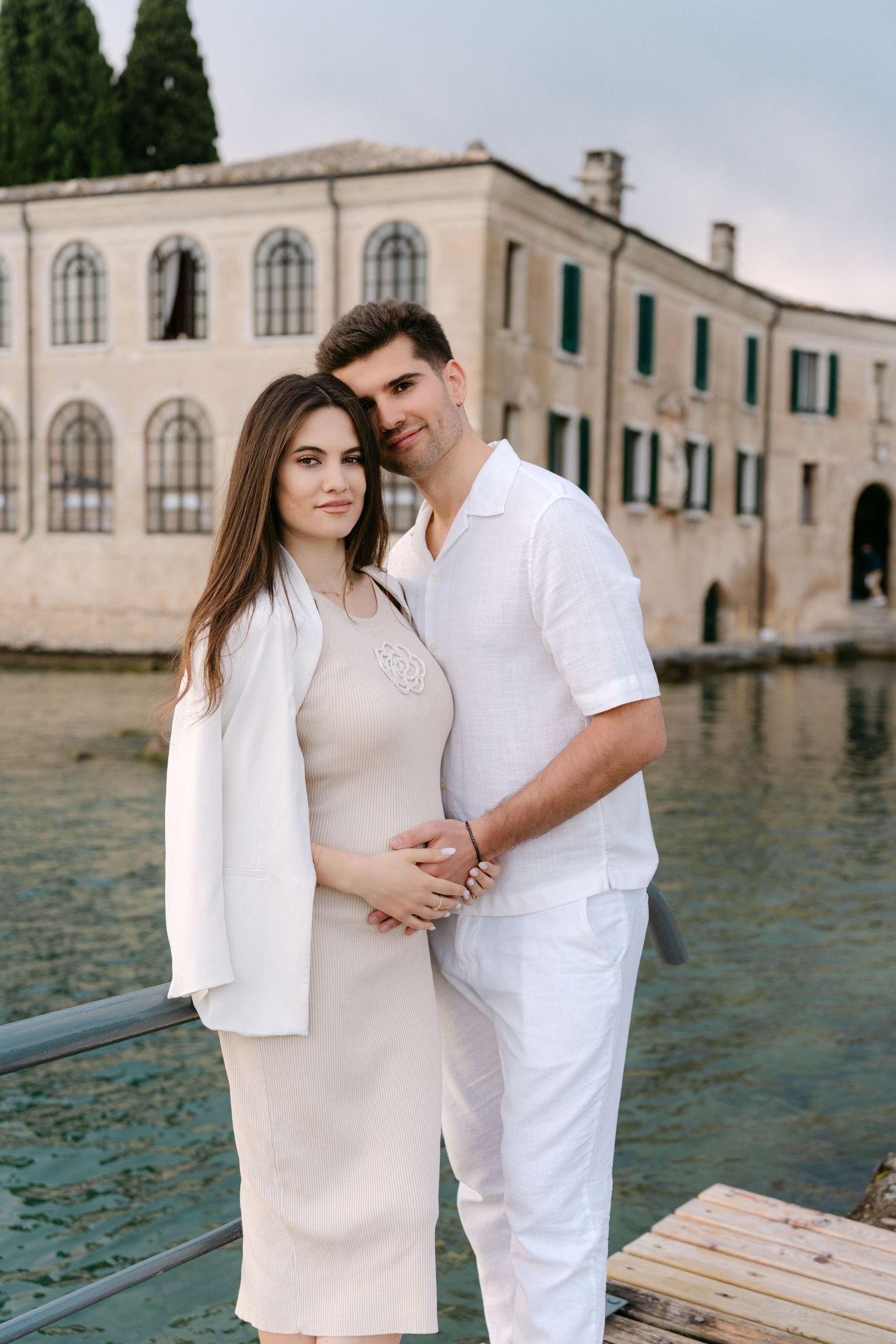 A couple in white outfits poses romantically by the water with historic architecture in the background.