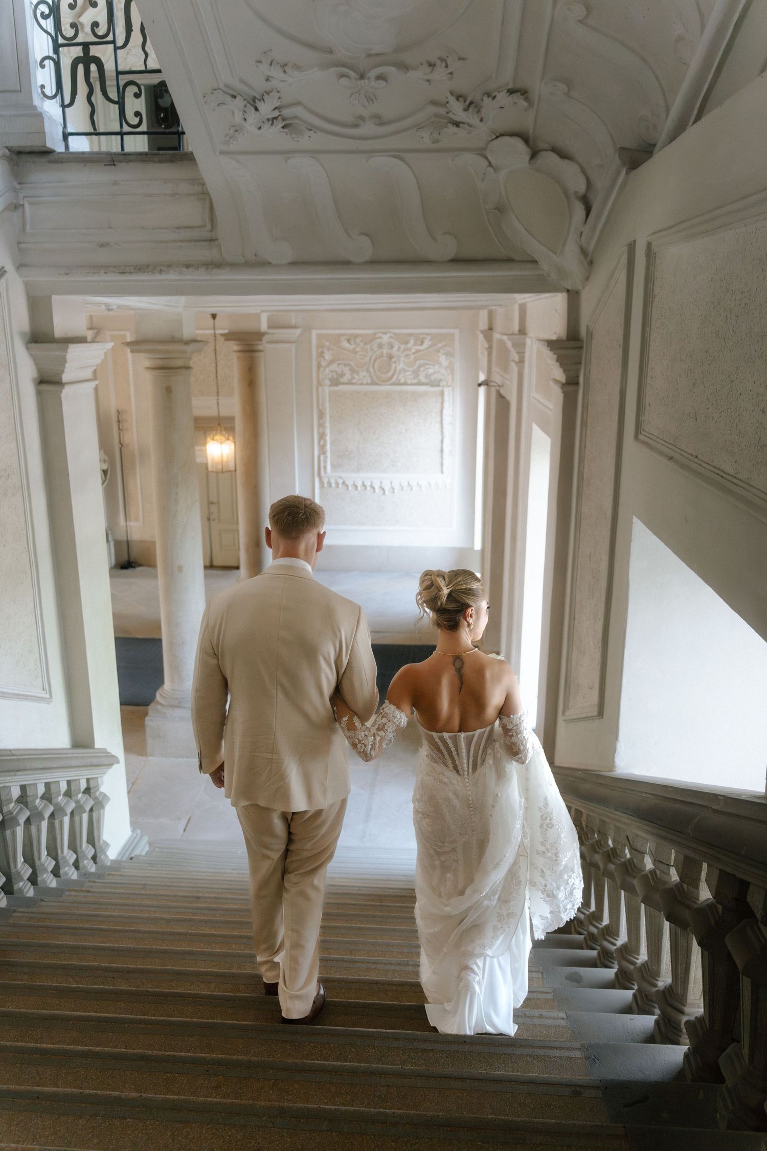 A couple in white wedding attire walks up an elegant marble staircase in a historic building. Schloss Ludwigsburg