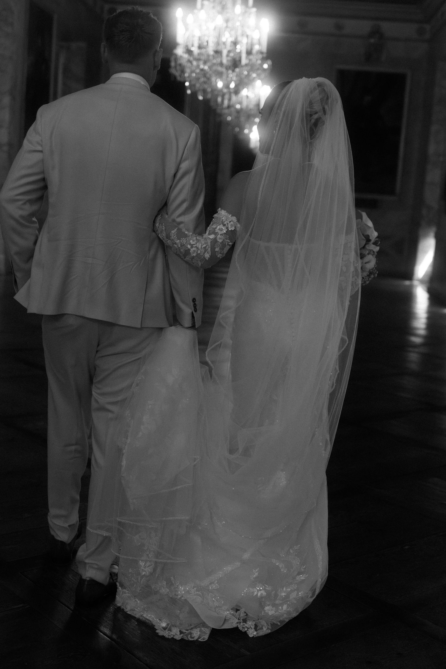 A couple in wedding attire walks through a dimly lit ballroom with crystal chandeliers overhead. Schloss Ludwigsburg