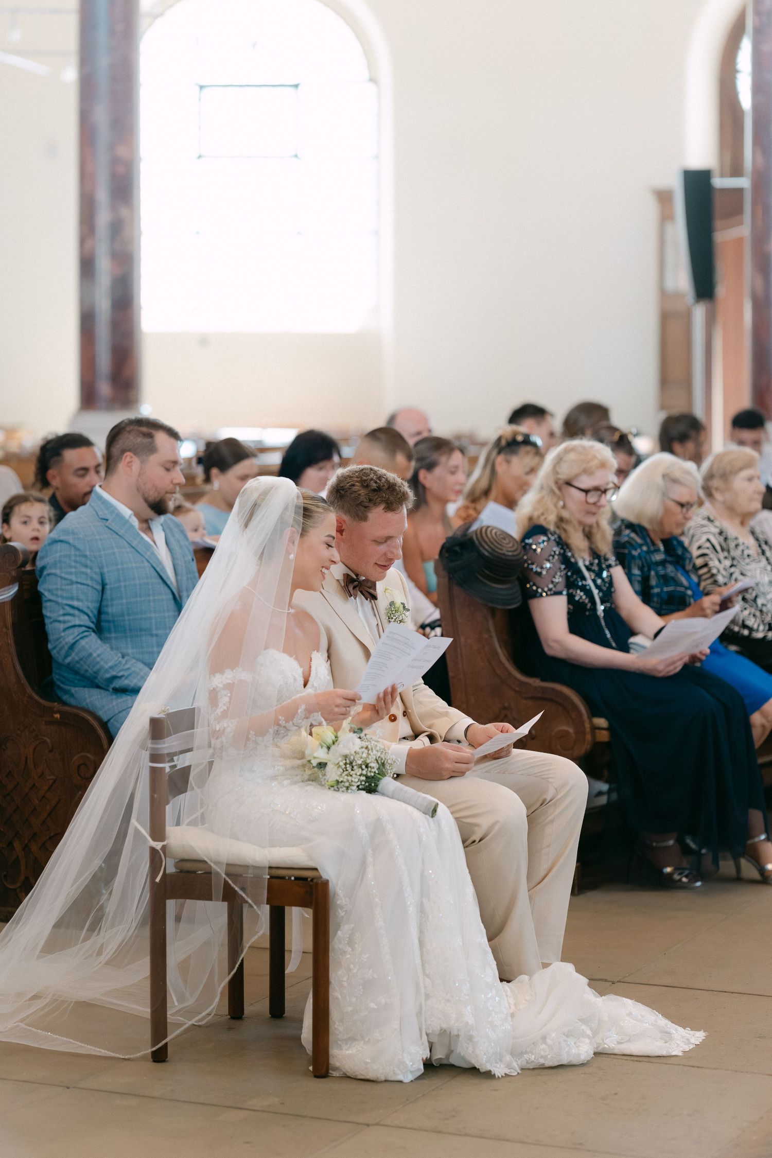 A bride in a white wedding dress sits with guests during an indoor church ceremony.