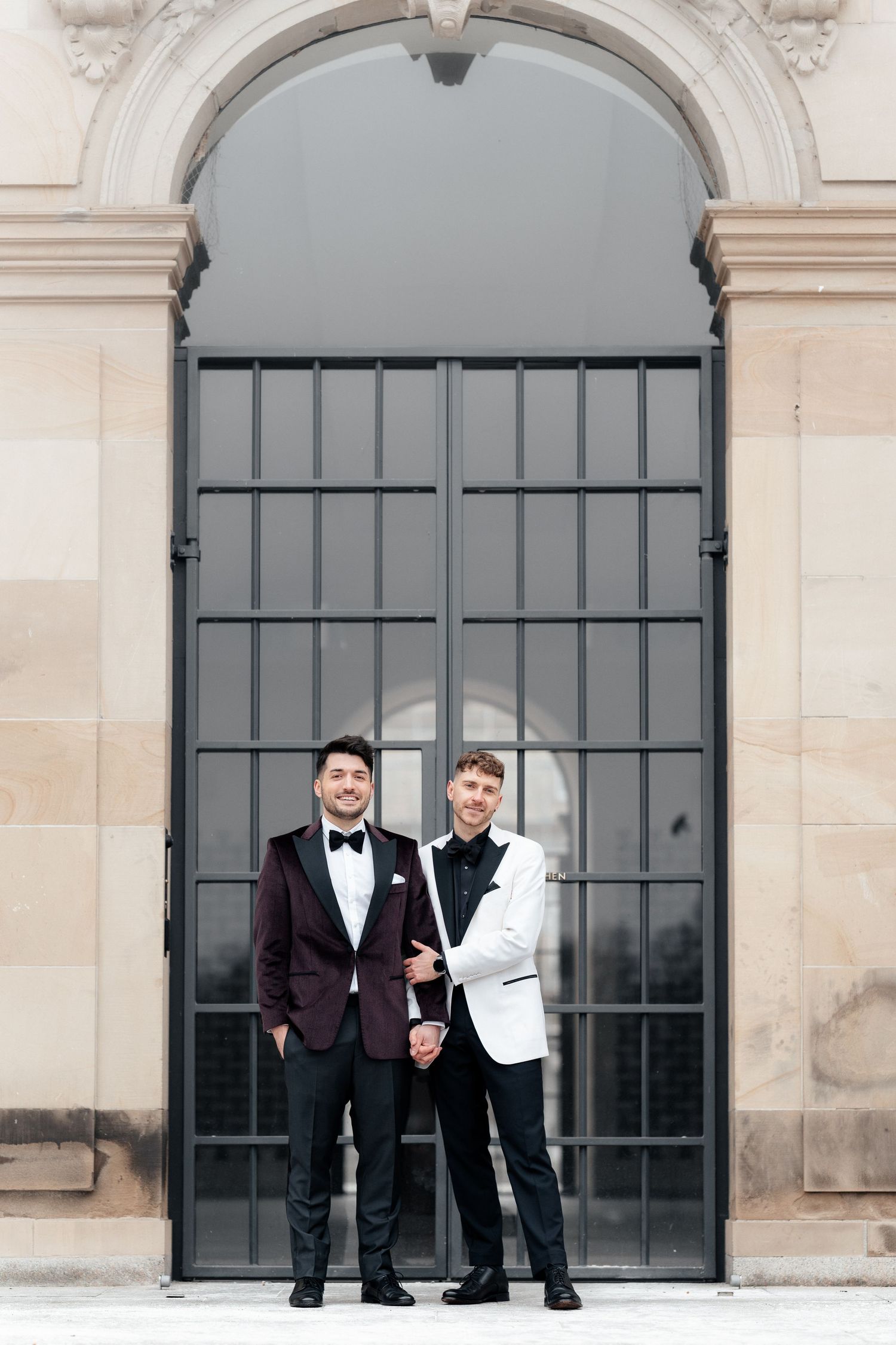 Two formally dressed people in tuxedos stand in front of a grand arched doorway with iron grillwork.